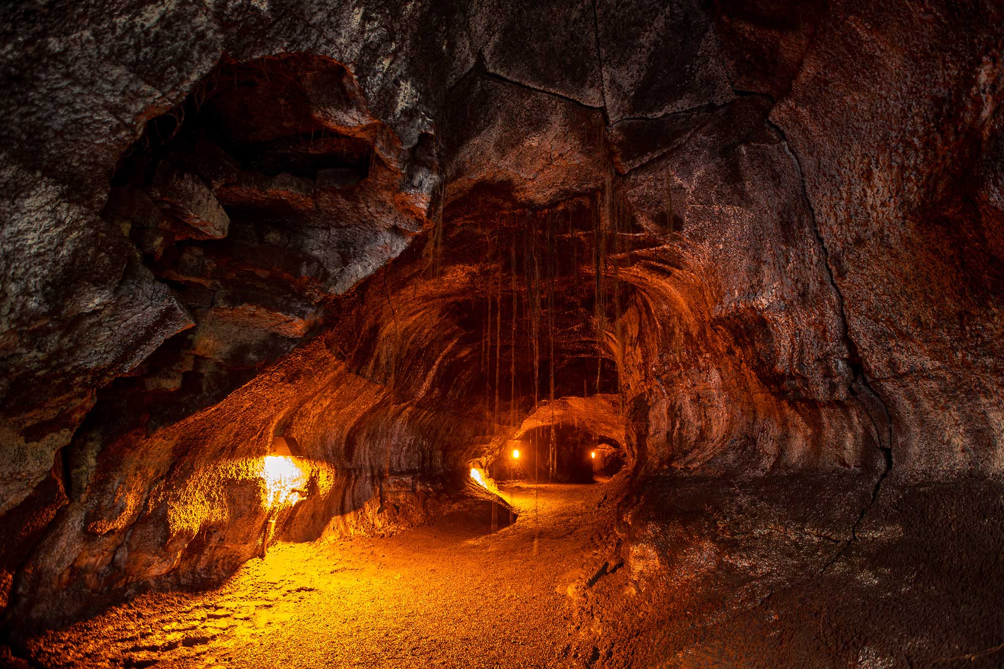 Photograph of the inside of Nahuku, a lava tube on Hawaii. The lava tube looks like a tunnel with a flat floor. Lights mounted in the tube illuminate the scene and small tree roots hang from the ceiling.