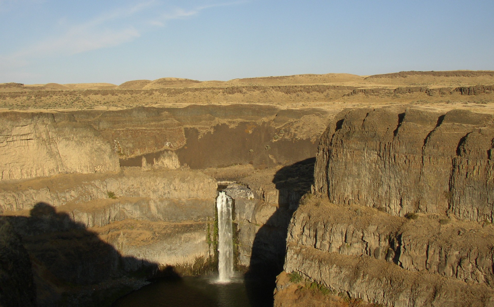 Photograph of Palouse Falls in eastern Washington. Palouse Falls is a tall waterfall that is falling straight down in a canyon. Steep cliffs with occasional ledges form the sides of the canyon. The landscape appears very dry with yellow vegetation. 
