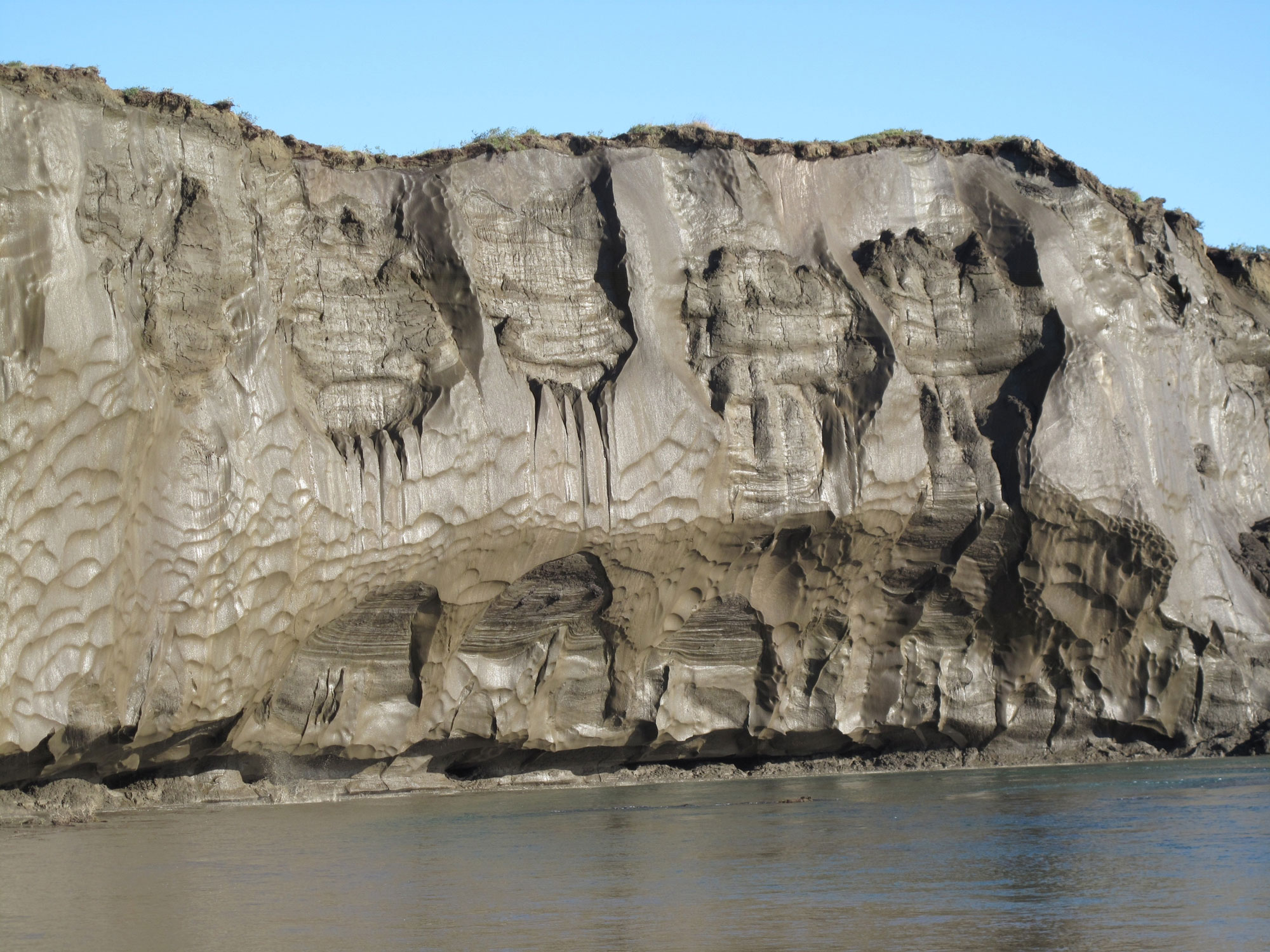 Photograph of permafrost exposed on a riverbank in Alaska. The photo shows a sheer bluff made up of brown ice with a rippled surface. A river can be seen at the base of the ice bluff.