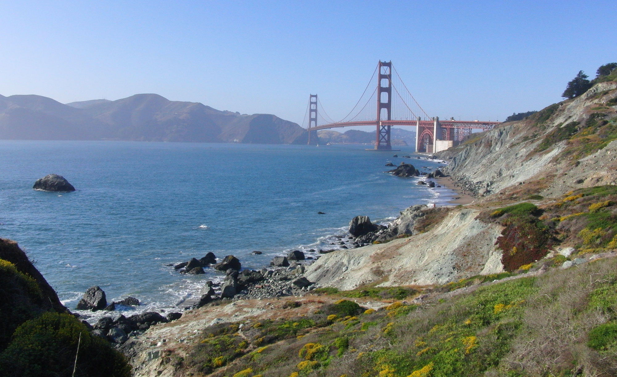 Photograph of bluffs by the side of the ocean in the Presidio, a park in San Francisco near the Golden Gate Bridge. In the foreground, greenish bluffs slope toward blue water. In the background, the Golden Gate Bridge spans the entrance to San Francisco Bay.
