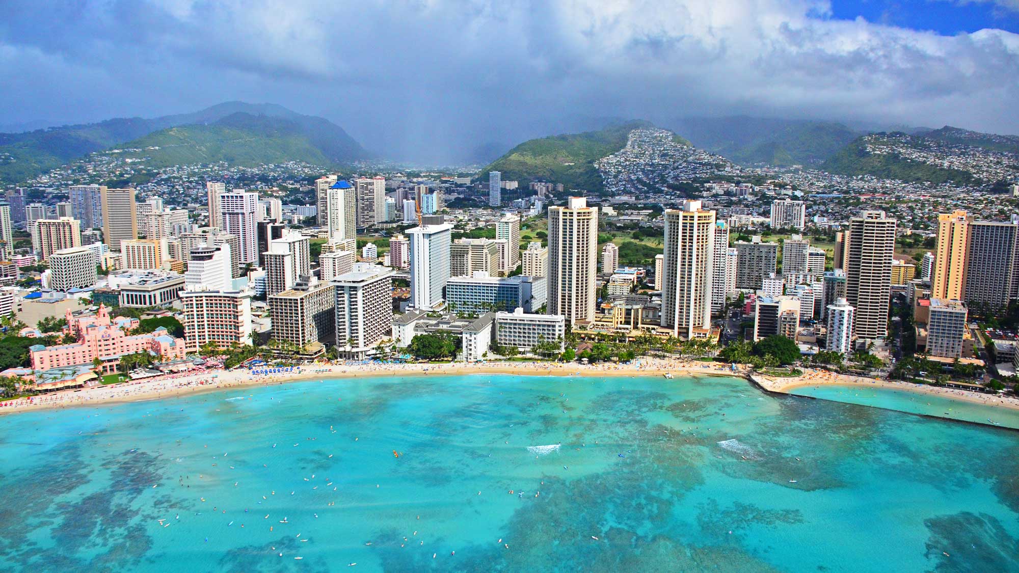 Photograph of Waikiki, Honolulu, O'ahu. The photo shows tall towers bordering a strip of white beach next of bright blue water. In the background, building of a city stretch into the distance up vegetated hillsides.