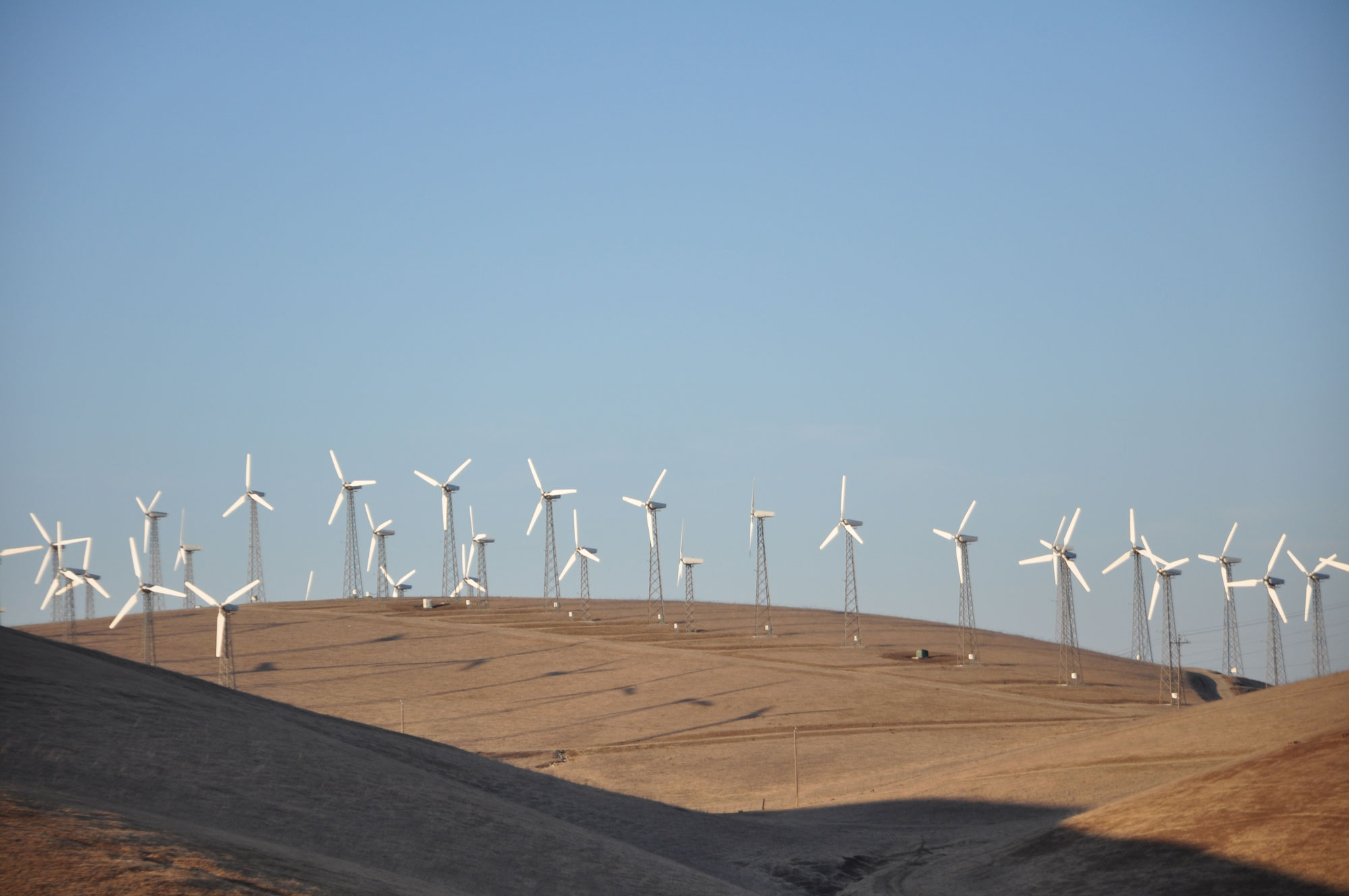 Photograph of the Altamont Wind Farm in California. The photo shows numerous white wind turbines dotting a landscape of rolling hills covered by yellow vegetation, probably grass.