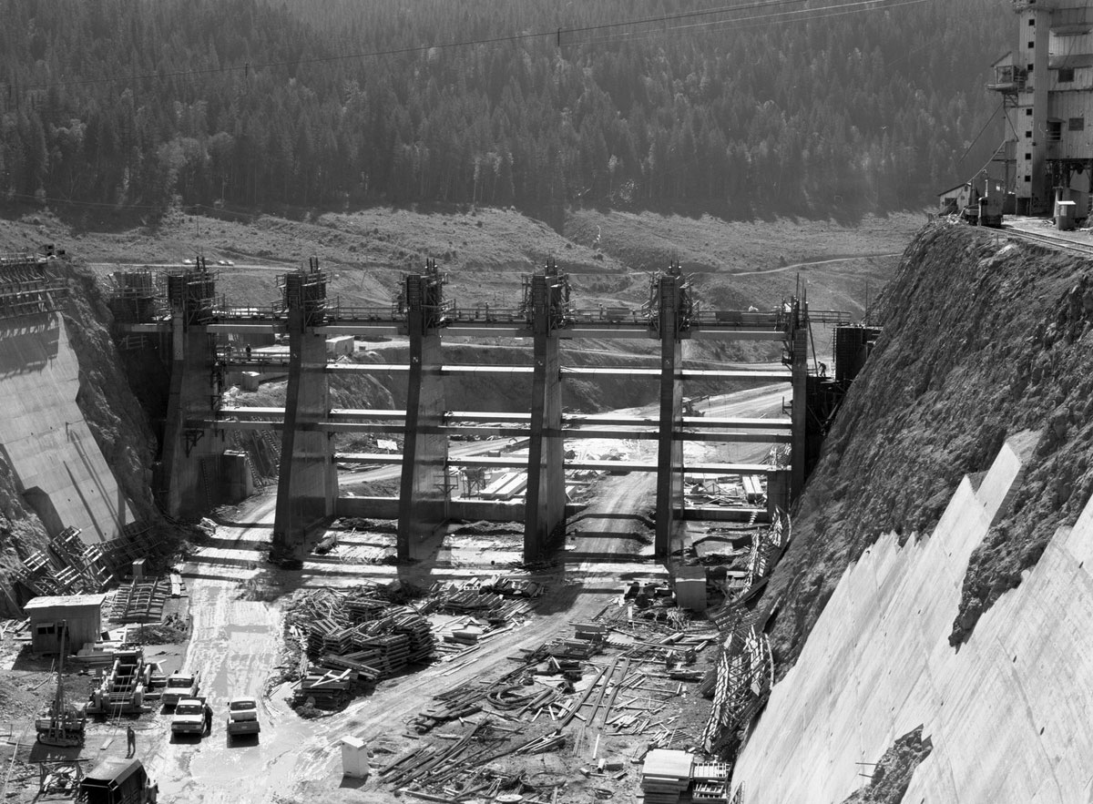 Black and white photograph of Boundary Dam under construction in northeastern Washington state in 1966. The photo shows the frame of the dam in place between two steeply sloping walls, one of concrete and one of rock. Trucks and building materials are on the ground at the base of the partially built dam.