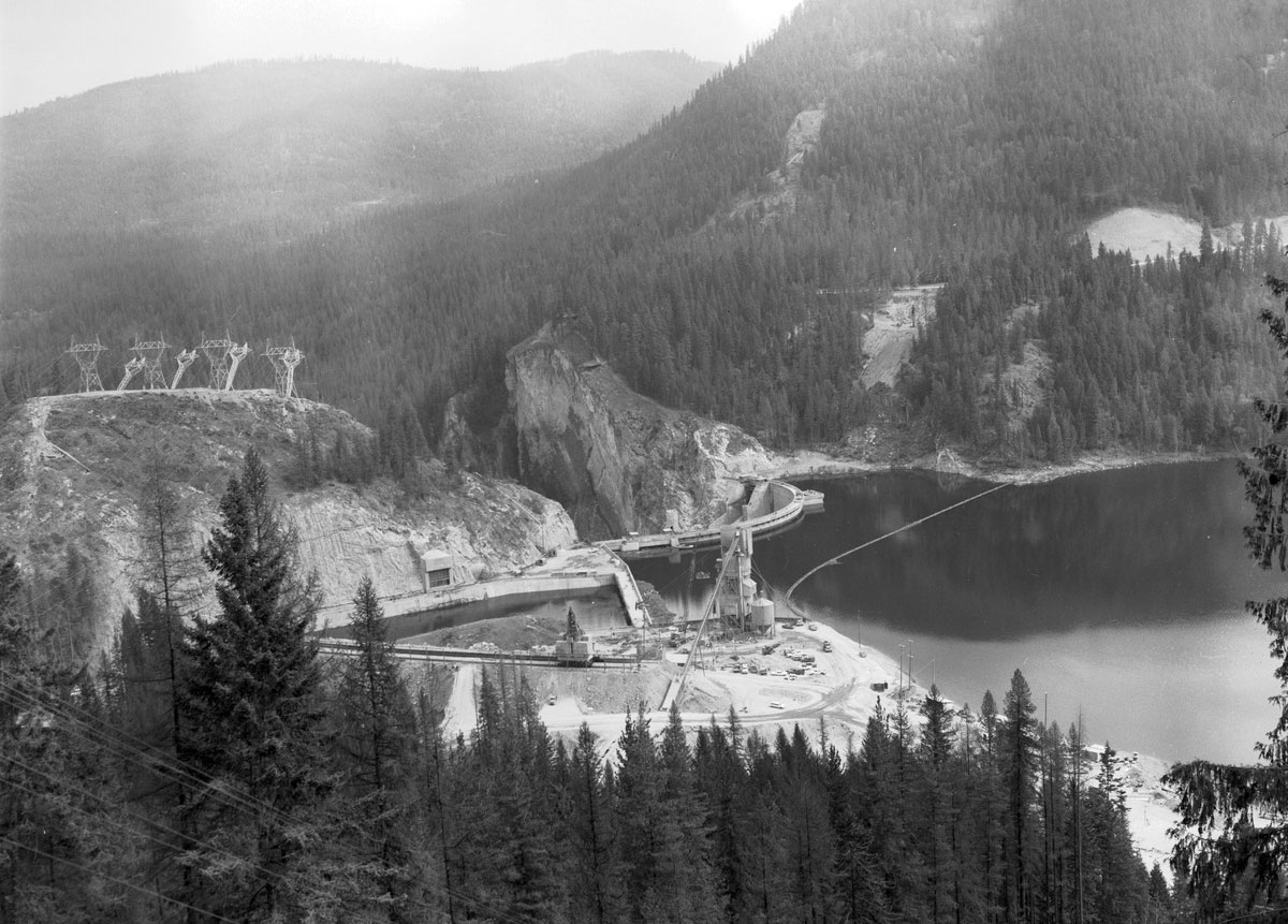 Black and white photograph of the Boundary Dam and Reservoir in Northeastern Washington state, 1967. The photo shows a reservoir on the right. The dam is in the center of the image, and the walls of a gorge are visible beyond it. Slopes covered with conifers surround the dam and reservoir.