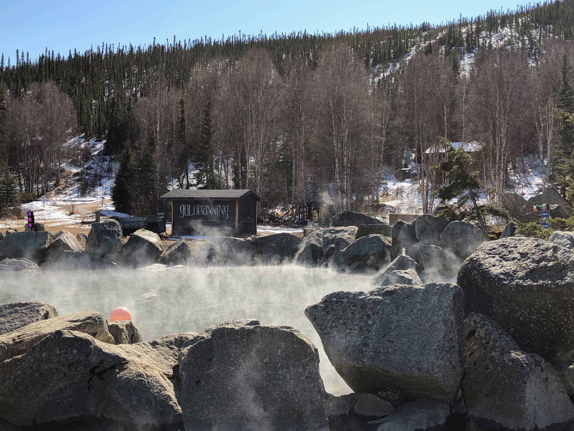 Photograph of a thermal pool at Chena Hot Springs, Alaska. The photo shows a pool of water surrounded by rocks. Steam hangs on the top of the pool. A hill covered with trees and snow rises in the background.