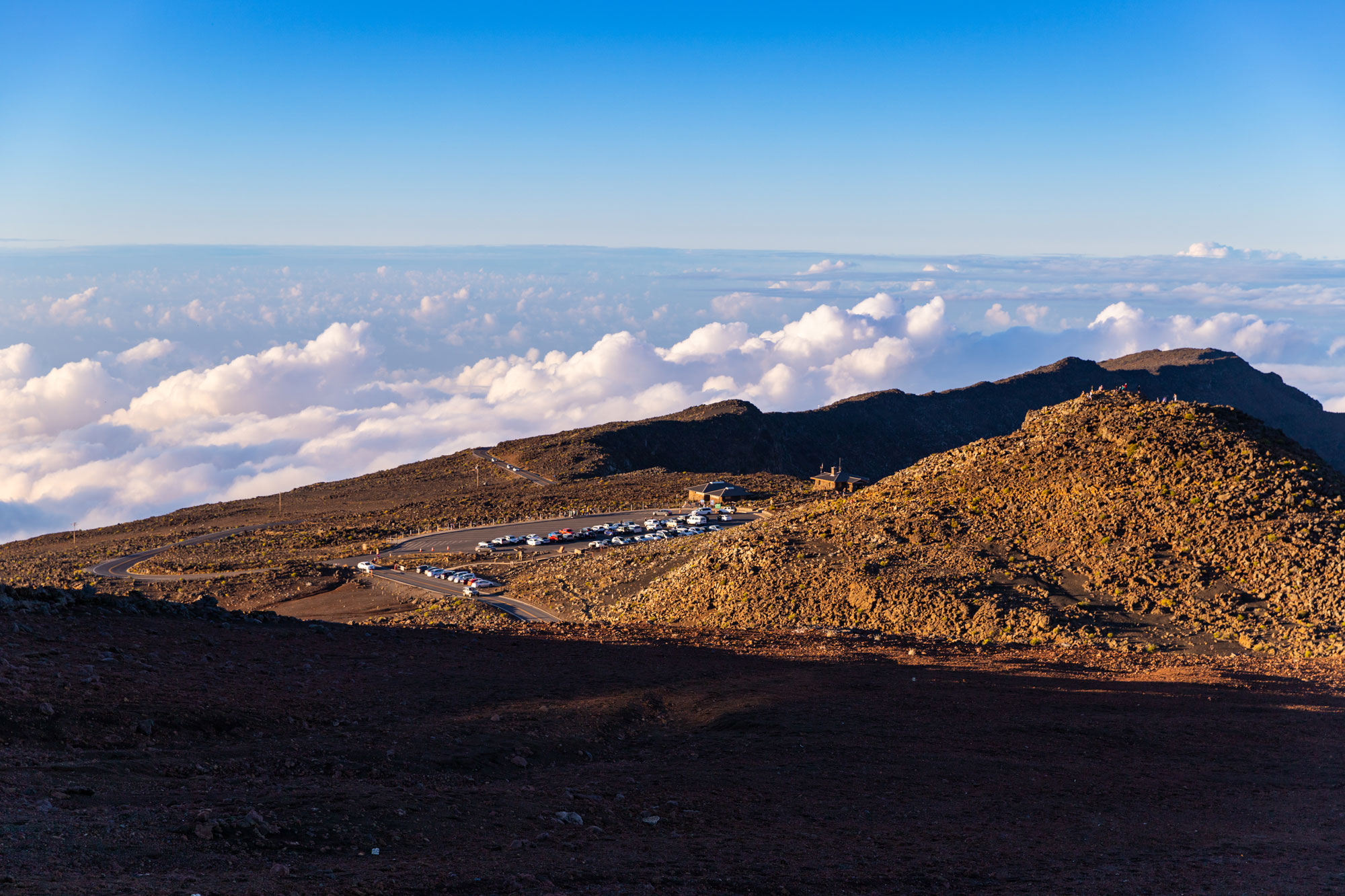 Photograph of the summit of Mount Haleakala on Maui. The photo shows a dry, barren beak with a parking area that has cars in it, next to which are some small buildings. Below the summit, clouds cover the sky, obscuring the ground below.