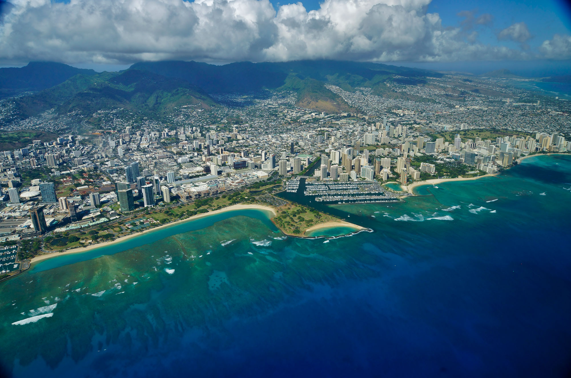 Aerial photograph of Waikiki, O'ahu, and nearby reefs. The photo shows building of a city, including many high-rises, next to the seashore. The coastline is fringed by a sandy beach. Wide coral reefs can be seen in the water just off the coast. They look like greenish and brownish patches in the water.