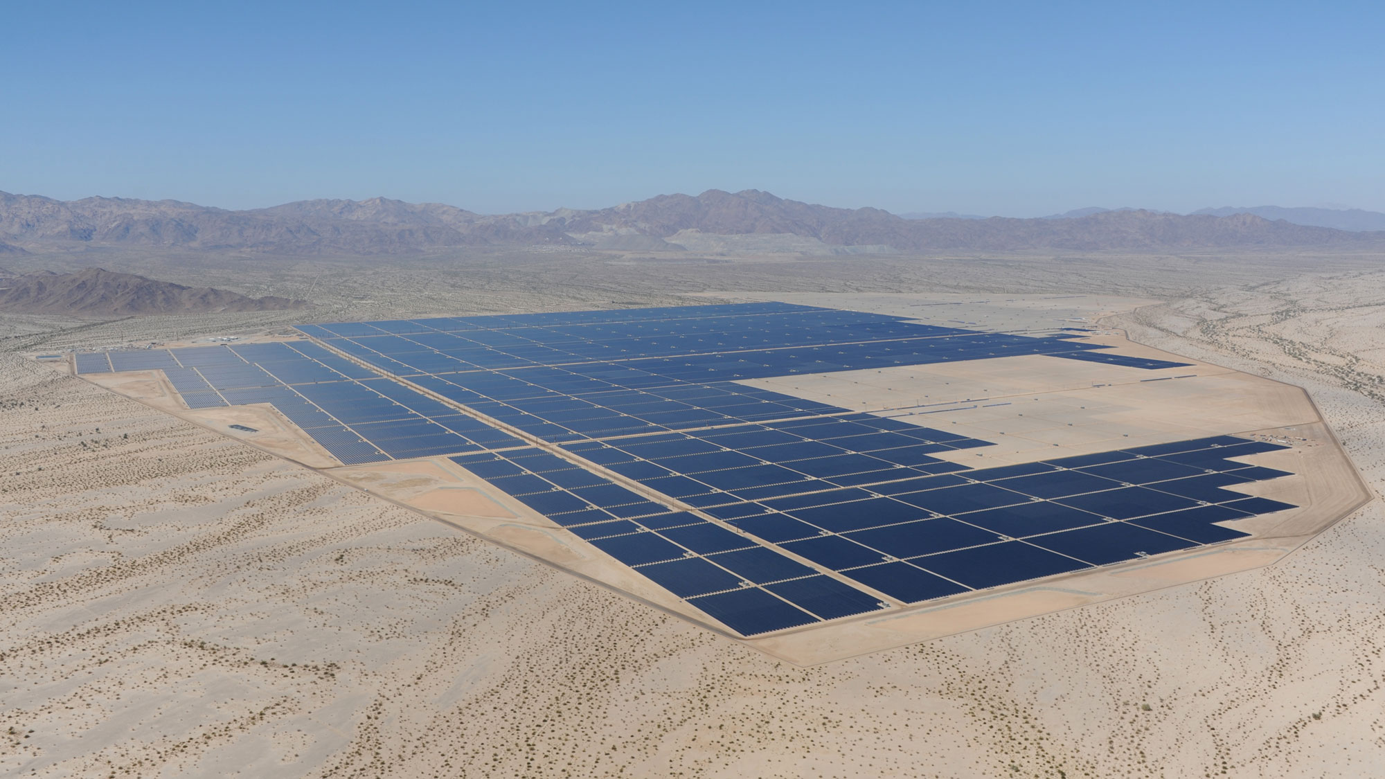 Aerial photograph of the Desert Sunlight Solar power plant in California. The photograph shows a series of black rectangles arrayed on a dry, relatively flat landscape. The rectangles are rows of solar panels seen from a distance. Mountains rise in the background.