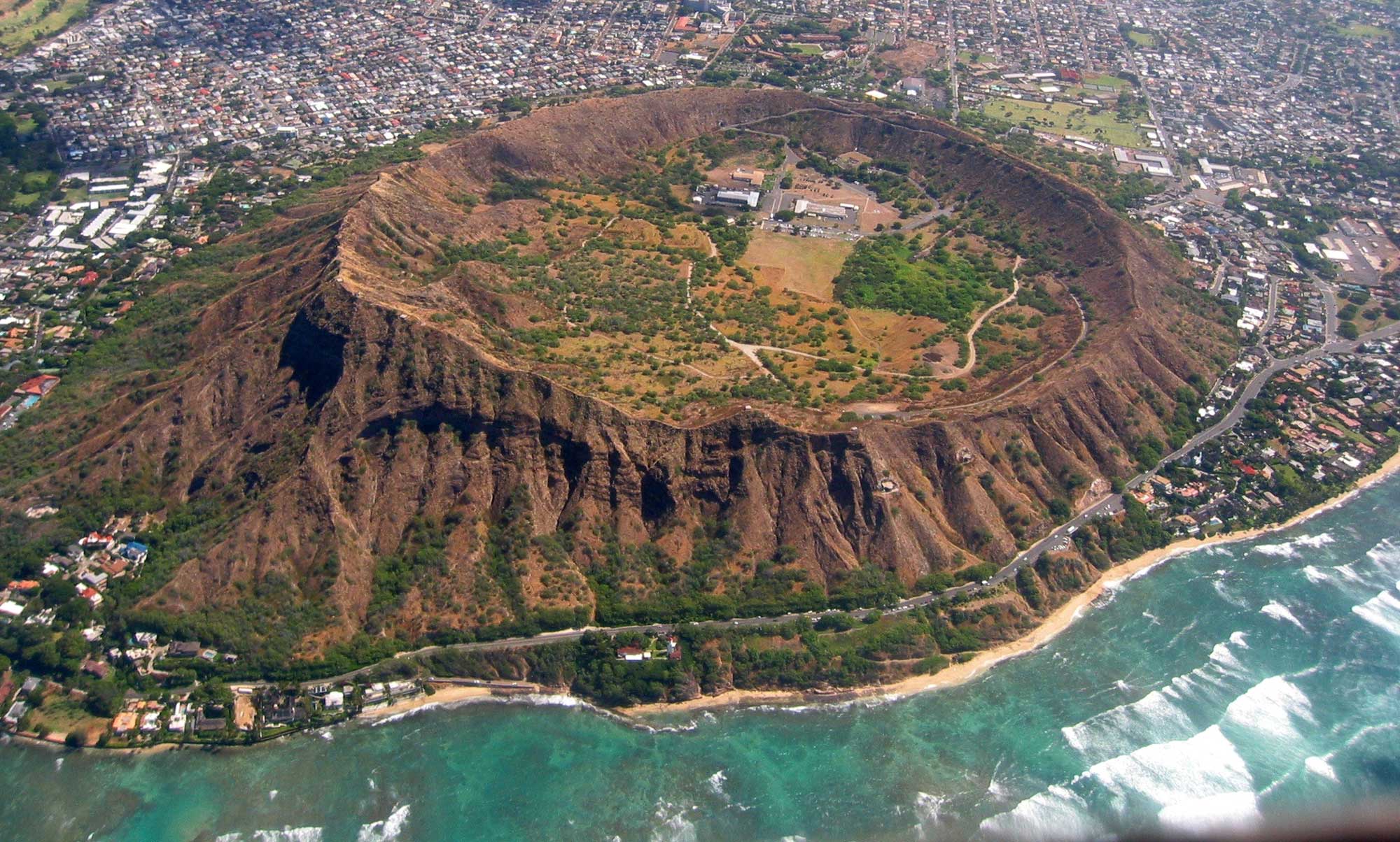 Photograph of Diamond Head, a large crater near the coast of Oahu. The photo shows a circular, raised crater with a narrow rim near the coast, with building of a city surrounding it on land. The bottom of the crater is flat inside with some green vegetation. A few buildings and parking lots are inside the far side of the crater.