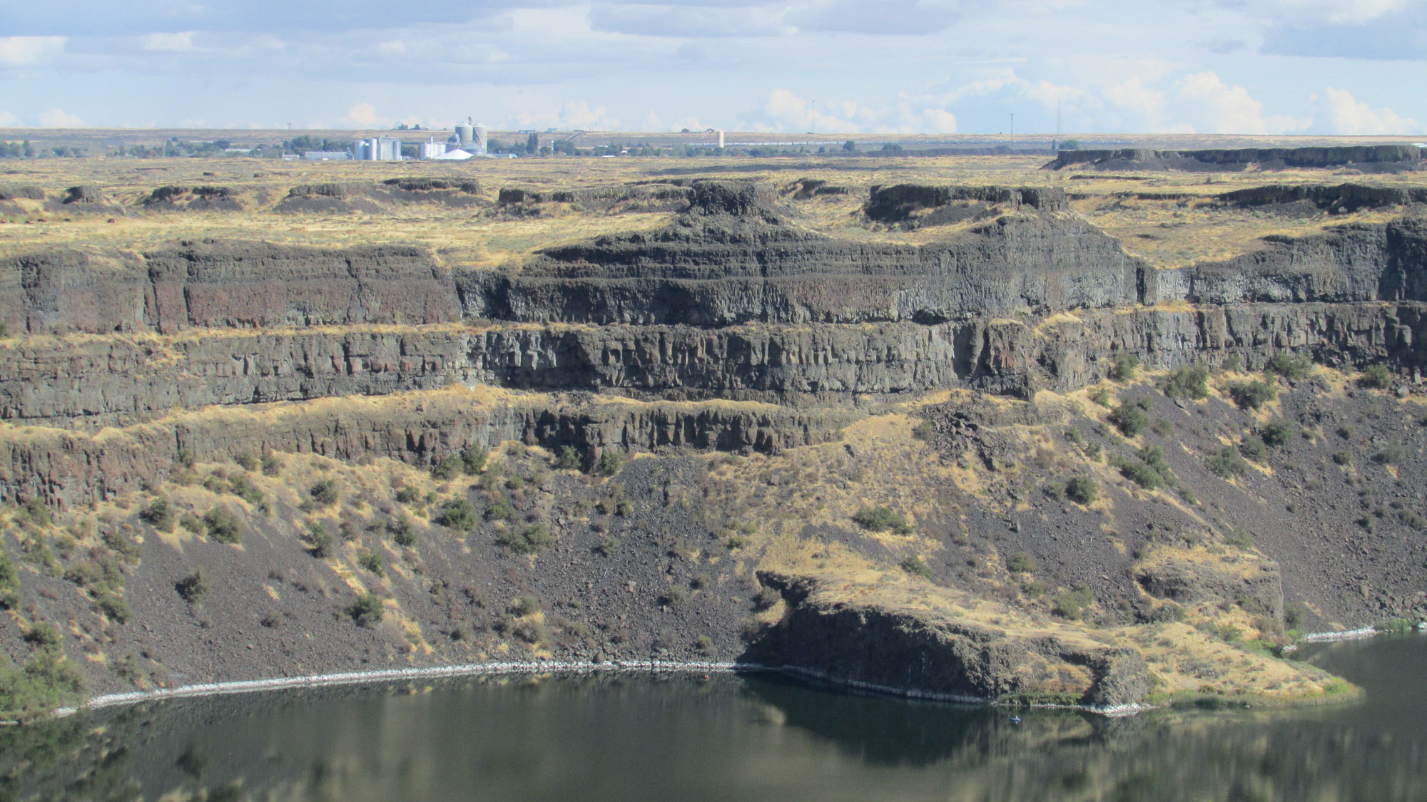 Photograph of basalt layers exposed surrounding Dry Falls Lake in Washington. The photo shows a tiered cliff at the edge of a lake. The cliff is made up of gray-brown rock with sparse yellow grass and green shrubs.