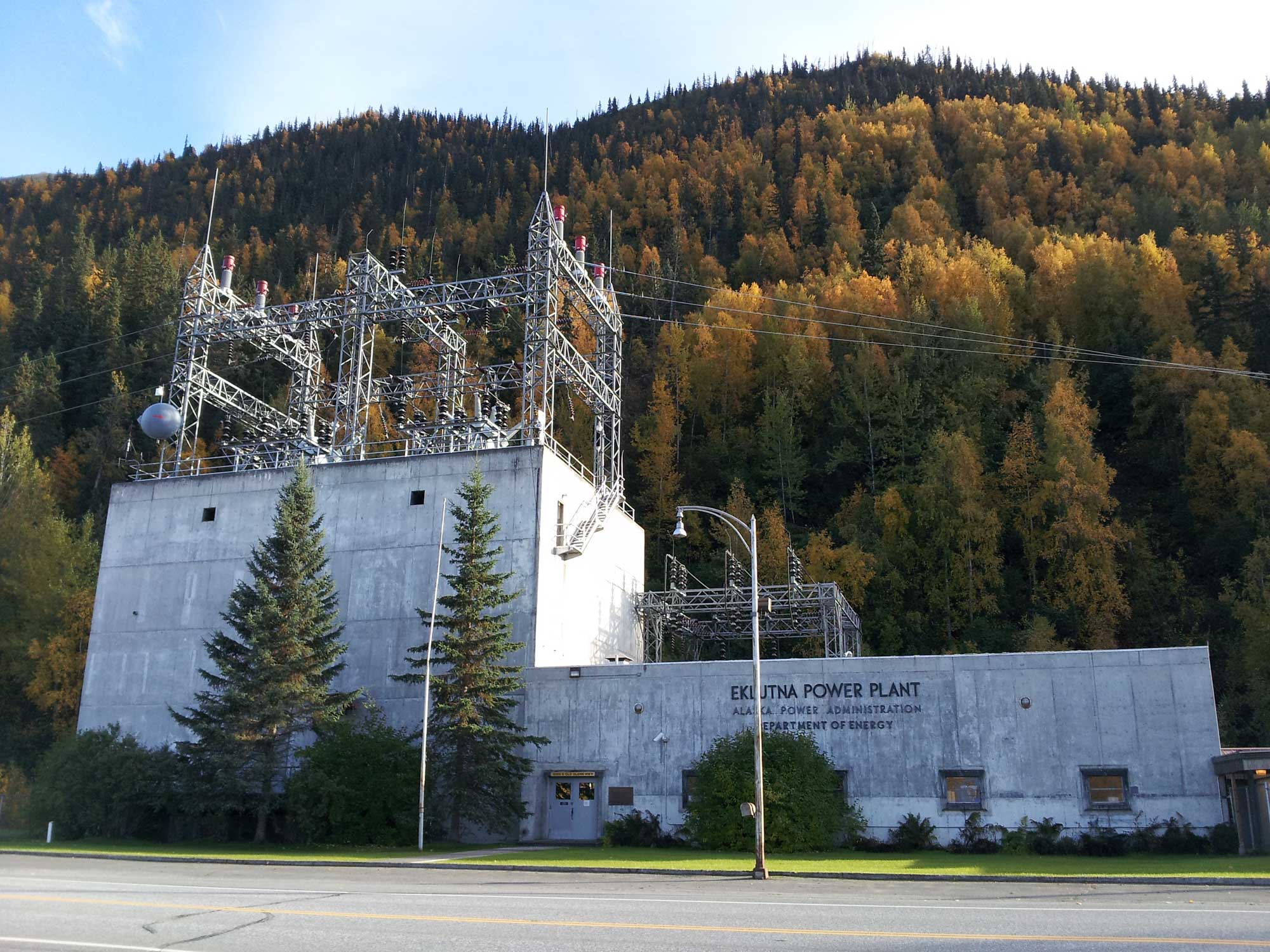 Photograph of the Eklunta Power Plant near Palmer, Alaska. The photo shows a concrete building with metal scaffolding on top to which power lines run. A hill covered with trees rises behind the plant.