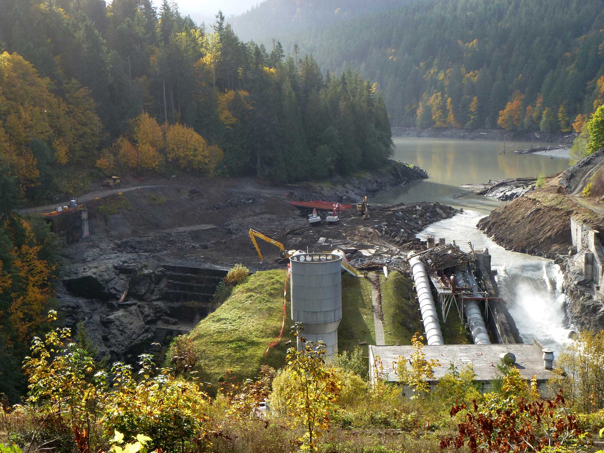 Photograph of the Elwha Dam removal on the Elwha River in Washington in 2011. In this photo, the dam is mostly removed, although some of the structures associated with it remain. Water flows through the area where the breached dam once was. Some construction equipment can be seen near the former dam site.