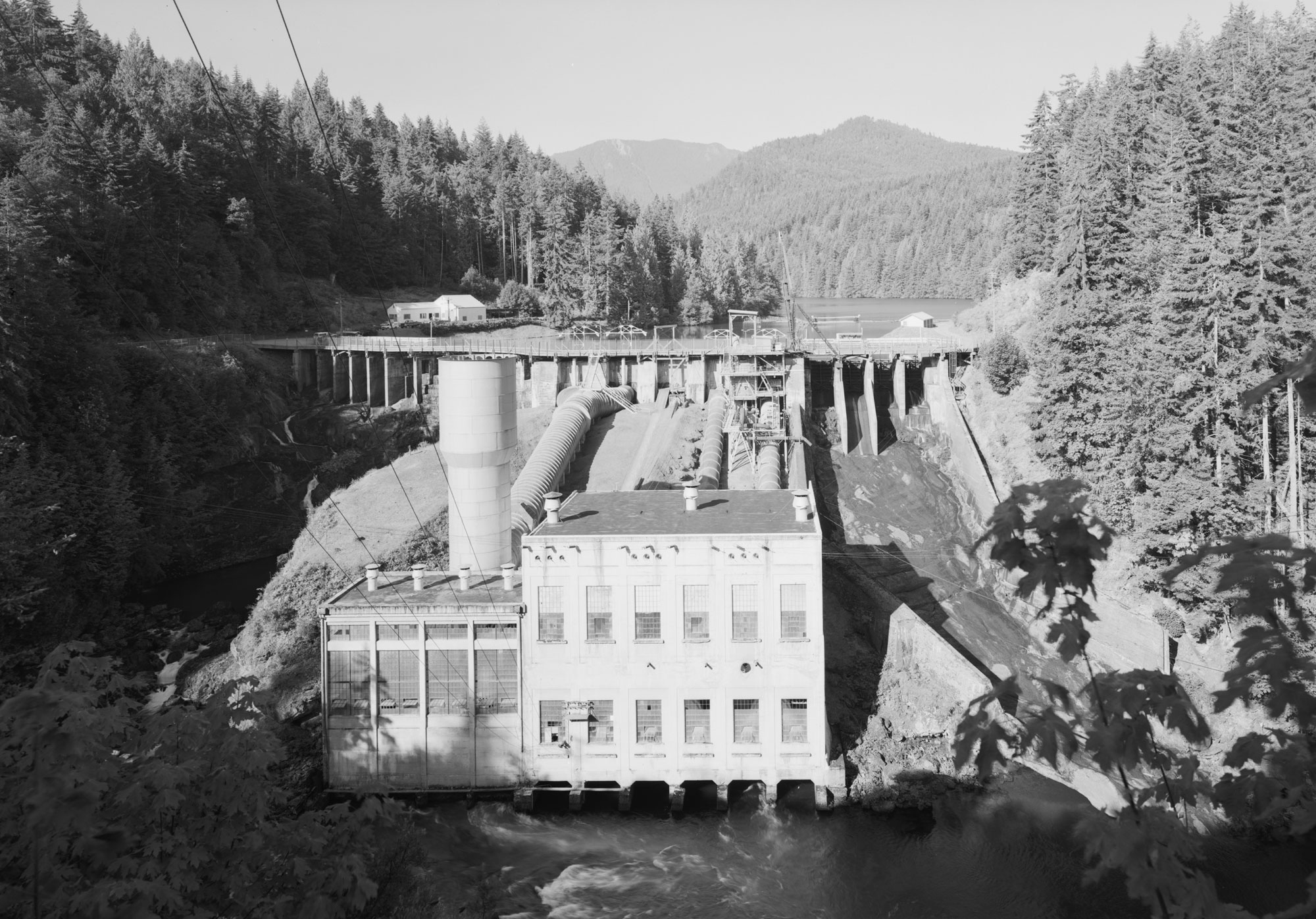 Black and white photograph of Elwha Dam and hydroelectric plant on the Elwha River in 1992. The photo shows the dam with a two-story building at its base. A cylindrical tower rises to the left of the building, beyond a shorter extension of the structure. A pipe runs from the dam to toward the building, the dam spillway it to the right. Hills covered with conifers arise around the dam and reservoir.