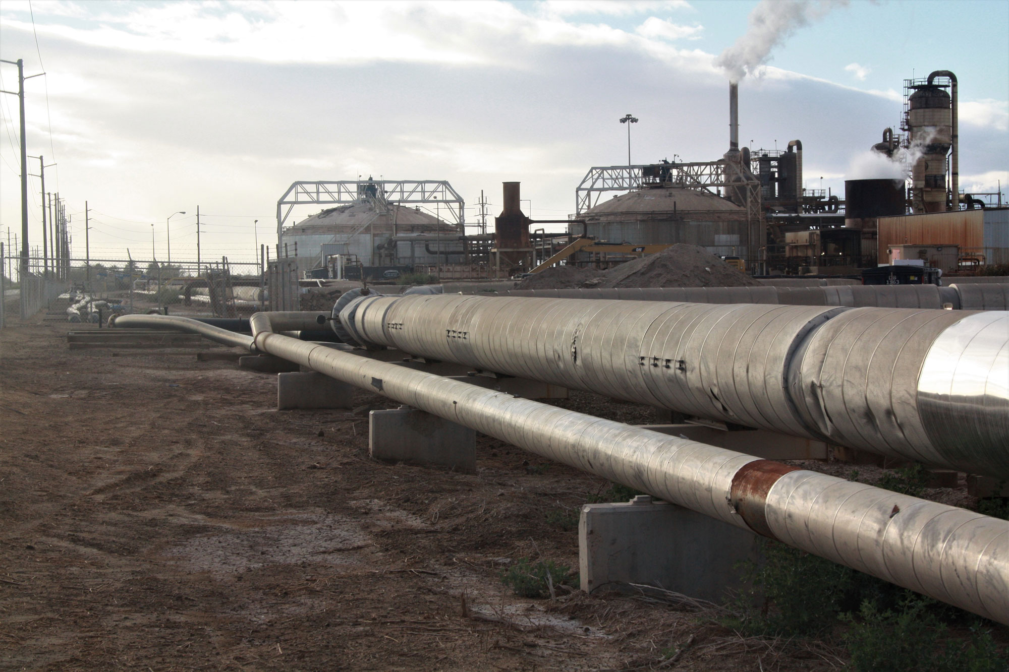 Photograph of part of a geothermal near the Salton Sea in southern California. The photo shows pipes in the foreground and cylindrical structures in the background, with steam rising from a smokestack.