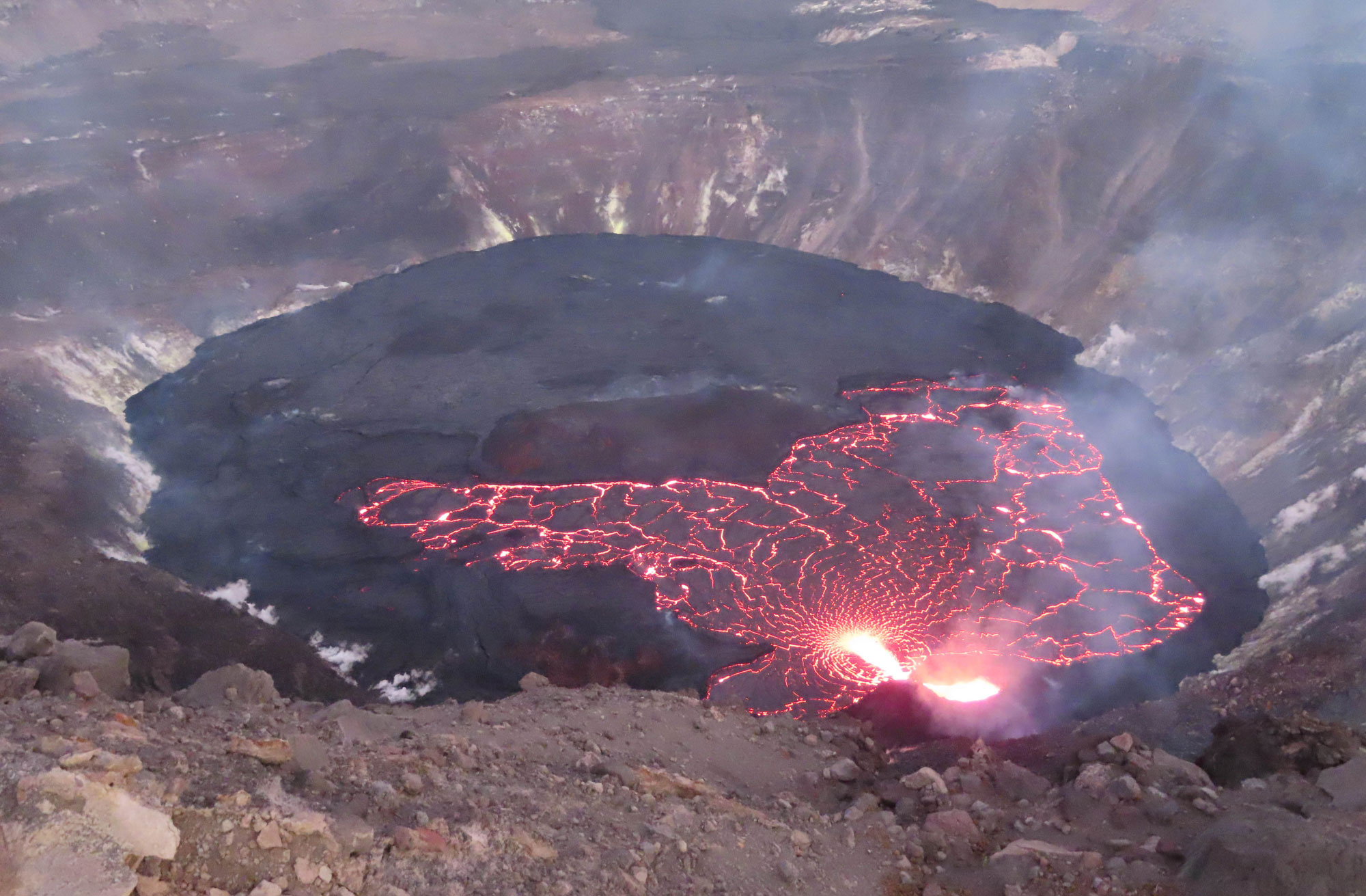 Photograph of a lava lake in Halema'uma'u crater, Kilauea caldera. The photos shows a round lava lake in a steep-sloped pit. The surface of the lake is a dull black color and partially cut by bright orange cracks formed by molten lava.