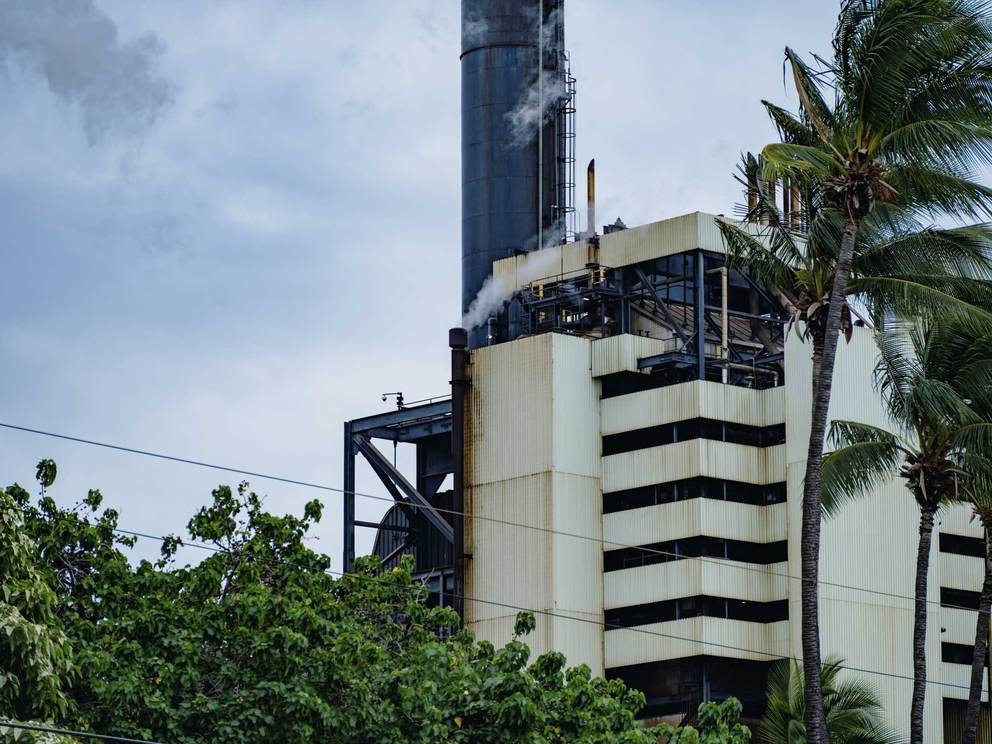 Photograph of a power plan in Kapolei, Oahu. The photo shows a white, multistory building rising behind treetops. A black smokestack rises behind the building, but the top of the smokestack is not visible.