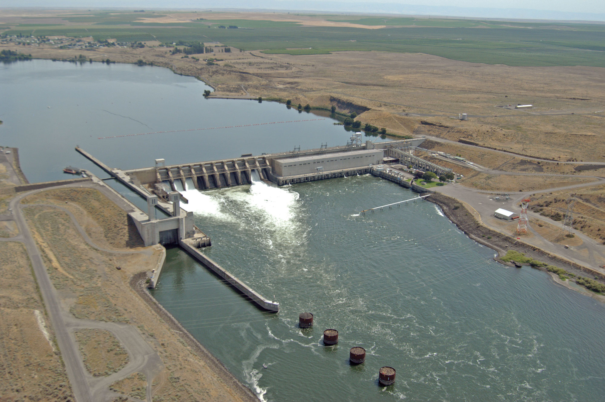 Aerial photograph of Ice Harbor Dam on the Snake River in eastern Washington. The photo shows a wide river with a concrete dam spanning it. The surrounding landscape is dry with yellow vegetation, although a large patch of green vegetation can be seen in the background.