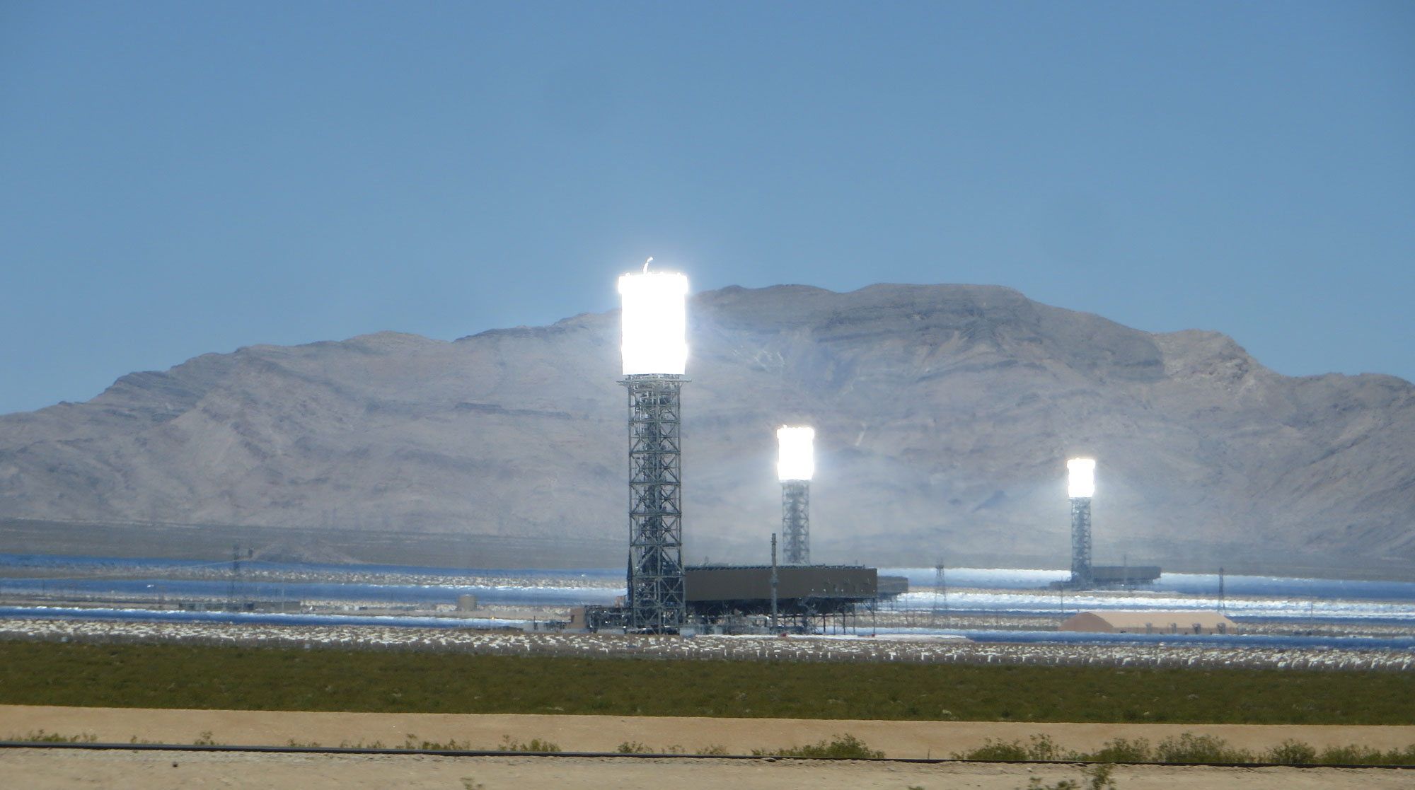 Ground-level photograph of the Ivanpah Solar Power Plant in California. The photo shows three towers made up of metal scaffolding, each with a cylindrical light at the top. On the ground, mirrors can be seen surrounding the towers. A mountain rises in the background.