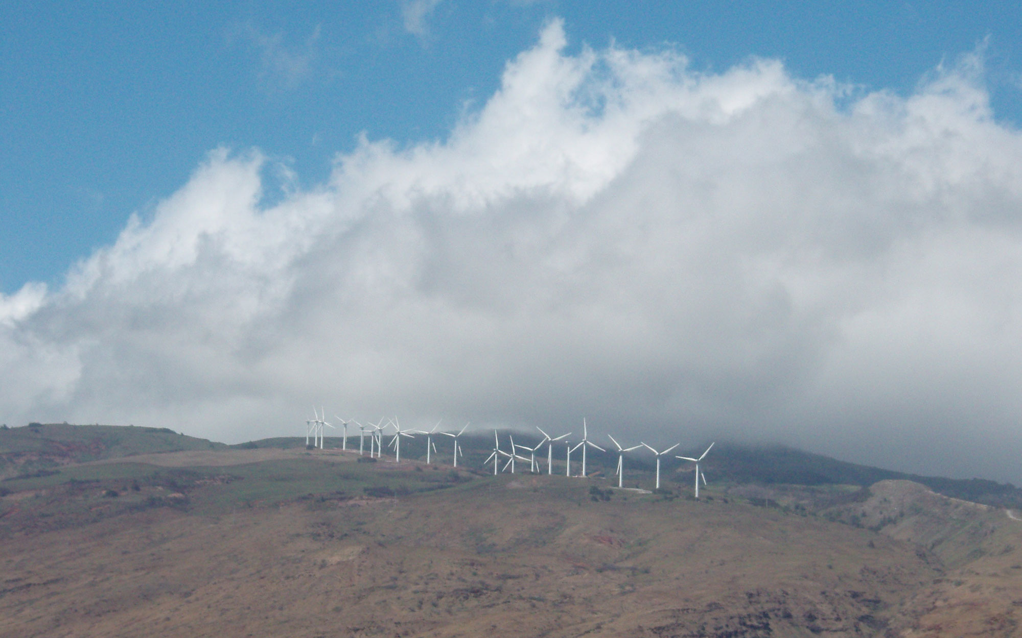 Photograph of Kaheawa wind farm, Maui, 2010. The photo shows a line of white wind turbines on a ridge with white clouds in the sky above. The ridge appears rather dry.