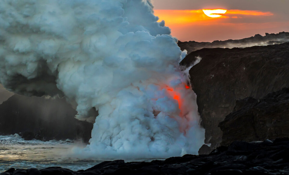 Photograph of lava flowing into the ocean on the coast of Hawai'i island at sunset. The photo shows steep, black lava-rock cliffs, a large plume of white steam coming from the ocean water, and an orange glove where lava is falling from the land.