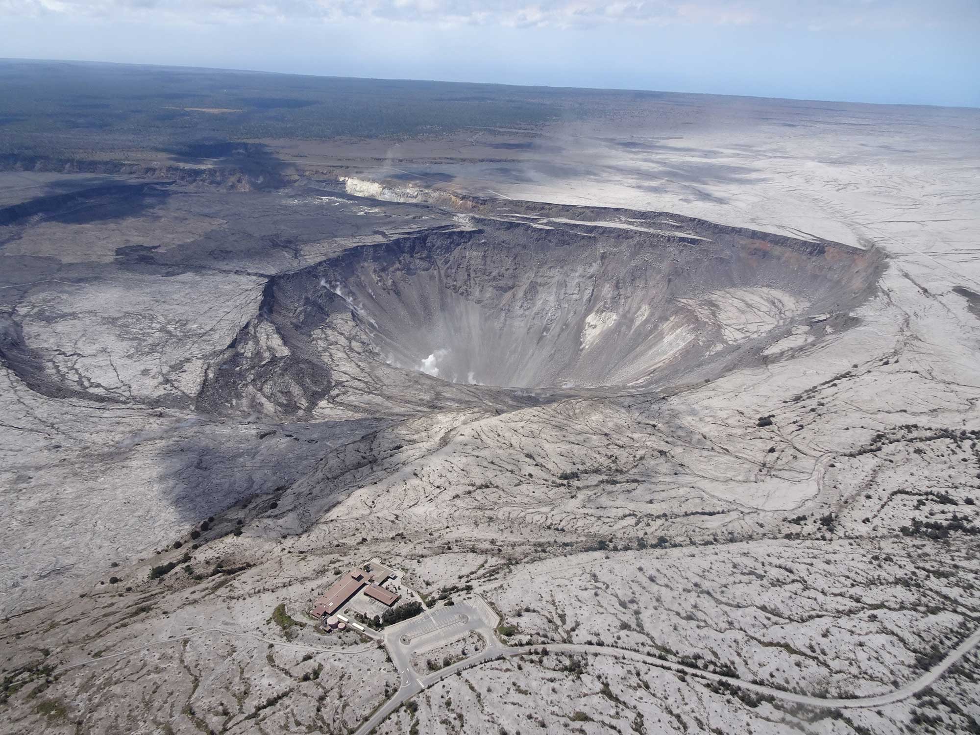 Photograph of the summit of Kilauea in July 2018 showing the caldera zone with Halema'uma'u crater appearing like a deep pit. The landscape around the crater is dry with sparse vegetation, and the Jagger Museum can be seen in the lower part of the image near the edge of the crater. The museum had to be closed in 2018 due to damage from earthquakes.