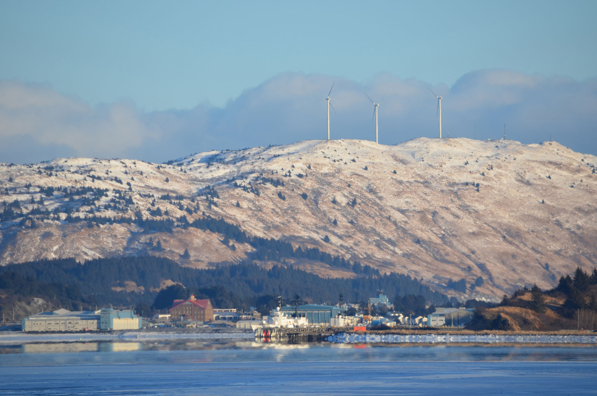 Photograph of wind turbines on Kodiak, Alaska. The photo shows three large white wind turbines on top of a snow-dusted hill. A cluster of buildings are grouped at the base of the hill, near a shoreline.
