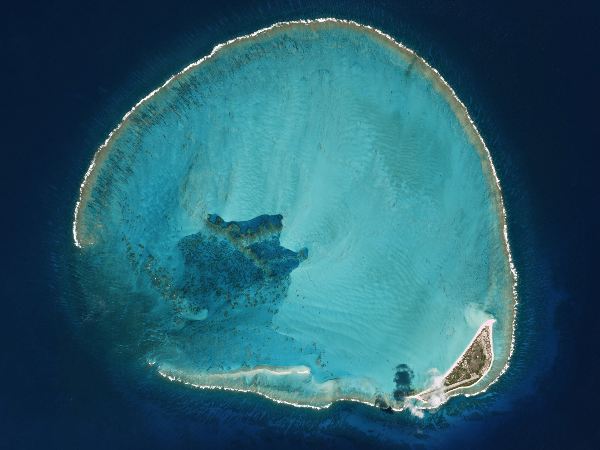 Photograph of Kure Atoll, probably from a satellite. The atoll looks like a thin white ring of land with dark water surrounding it and bright blue water within. In the lower right side of the Atoll is a larger piece of land with an airstrip.