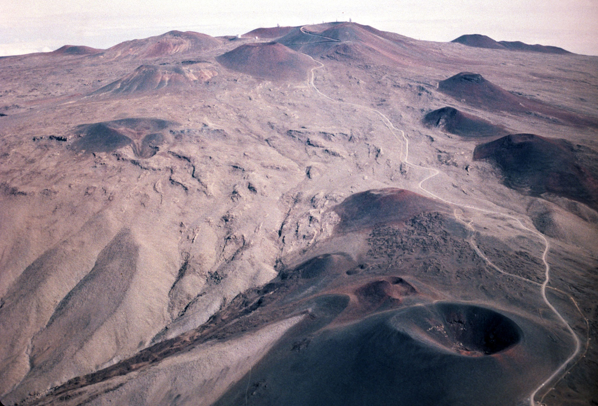 Photograph taken near the summit of Mauna Kea. Mauna Kea is in the post-caldera stage and the surface of the mountain near the summit has multiple cinder cones, giving it a "bumpy" appearance. The landscape is the photo is dry and lacks vegetation.