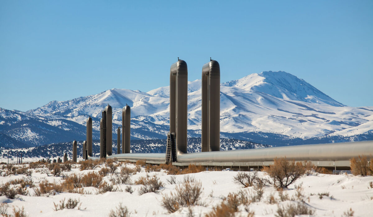 Photograph of geothermal pipes for the McGinness Hills geothermal plant in Nevada. The photo shows two parallel pipes running horizontally over a flat, snow-covered landscape. At regular intervals, each pipe has a vertical u-shaped bend. Mountains covered with snow rise in the background.