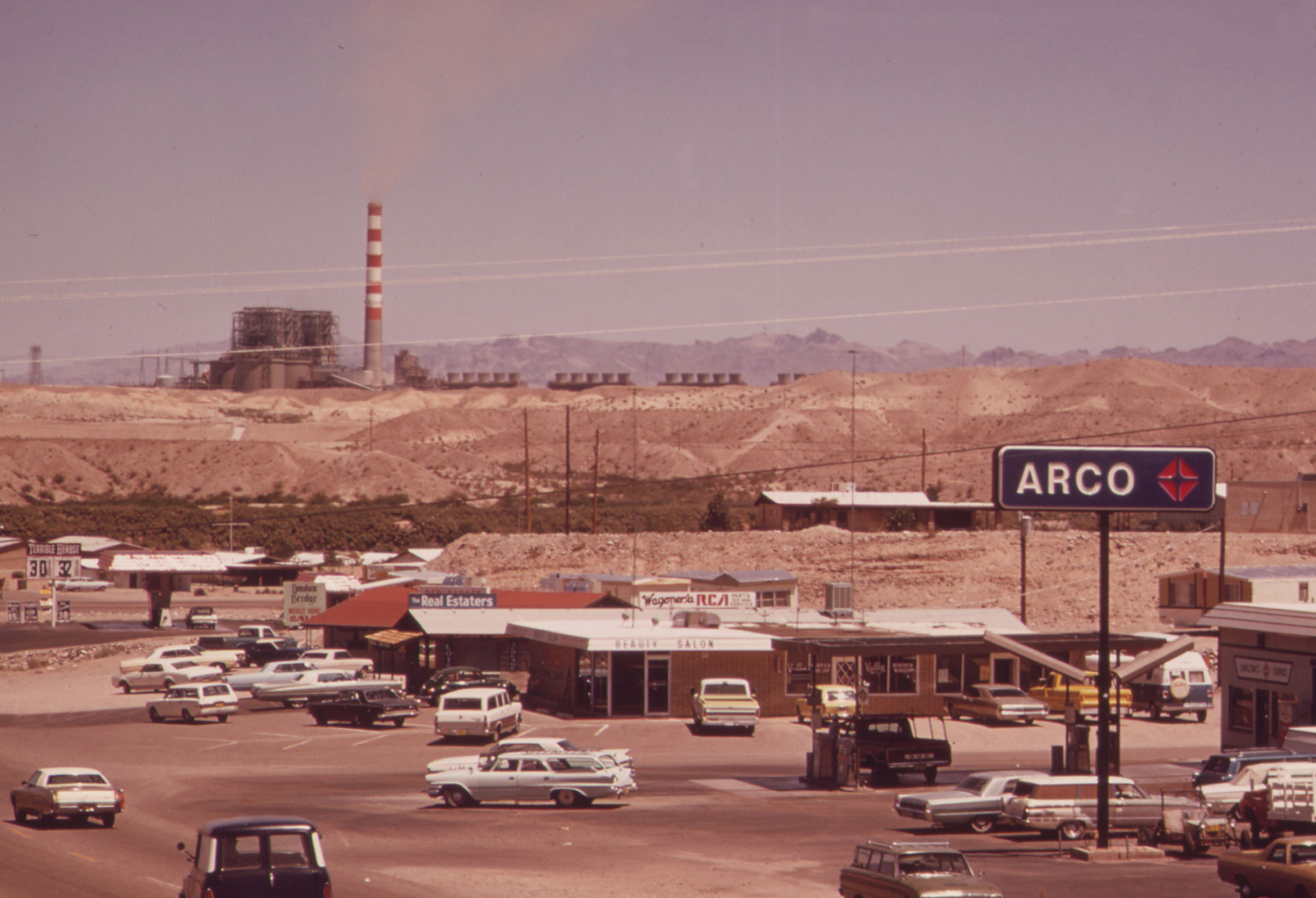Photograph of the Mohave Generating Station, a power plant that formally stood in Nevada. The photo shows a power station with a tall white-and-red striped smokestack on a hill in the background; other power plant structures are around it. In the foreground, old cars and buildings in Bullhead City, Arizona can be seen. The picture is from the 1970s.