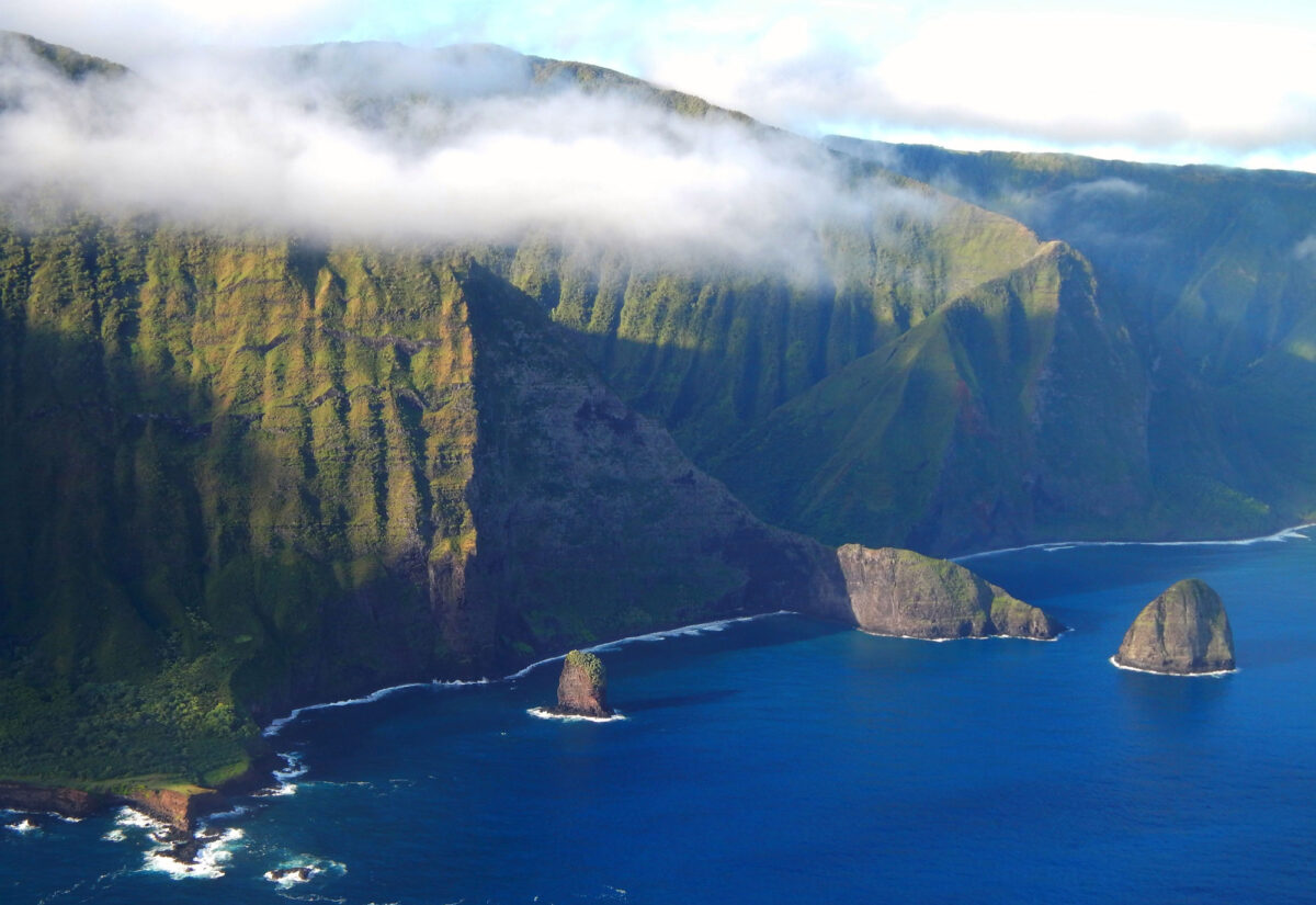 Photograph of steep cliffs on the shore of Molokai with a light layer of clouds near the tops. The ocean meets the bottoms of the cliffs, and two small islets stand in the water near them.