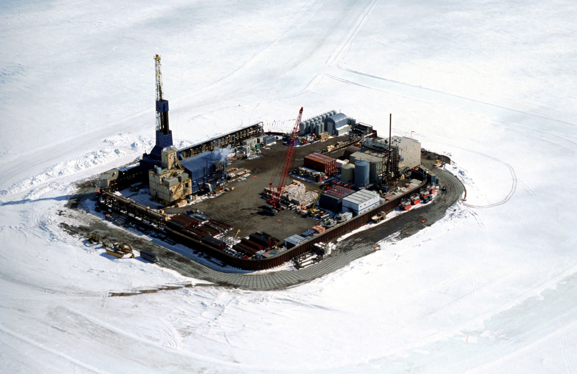 Aerial photograph of Northstar Island, a man-made island in the Beaufort Sea that supports oil extraction equipment. The photo shows a rectangular island with a drill rig and other equipment on it. The island is surrounded by sea ice covered in a thin layer of snow.
