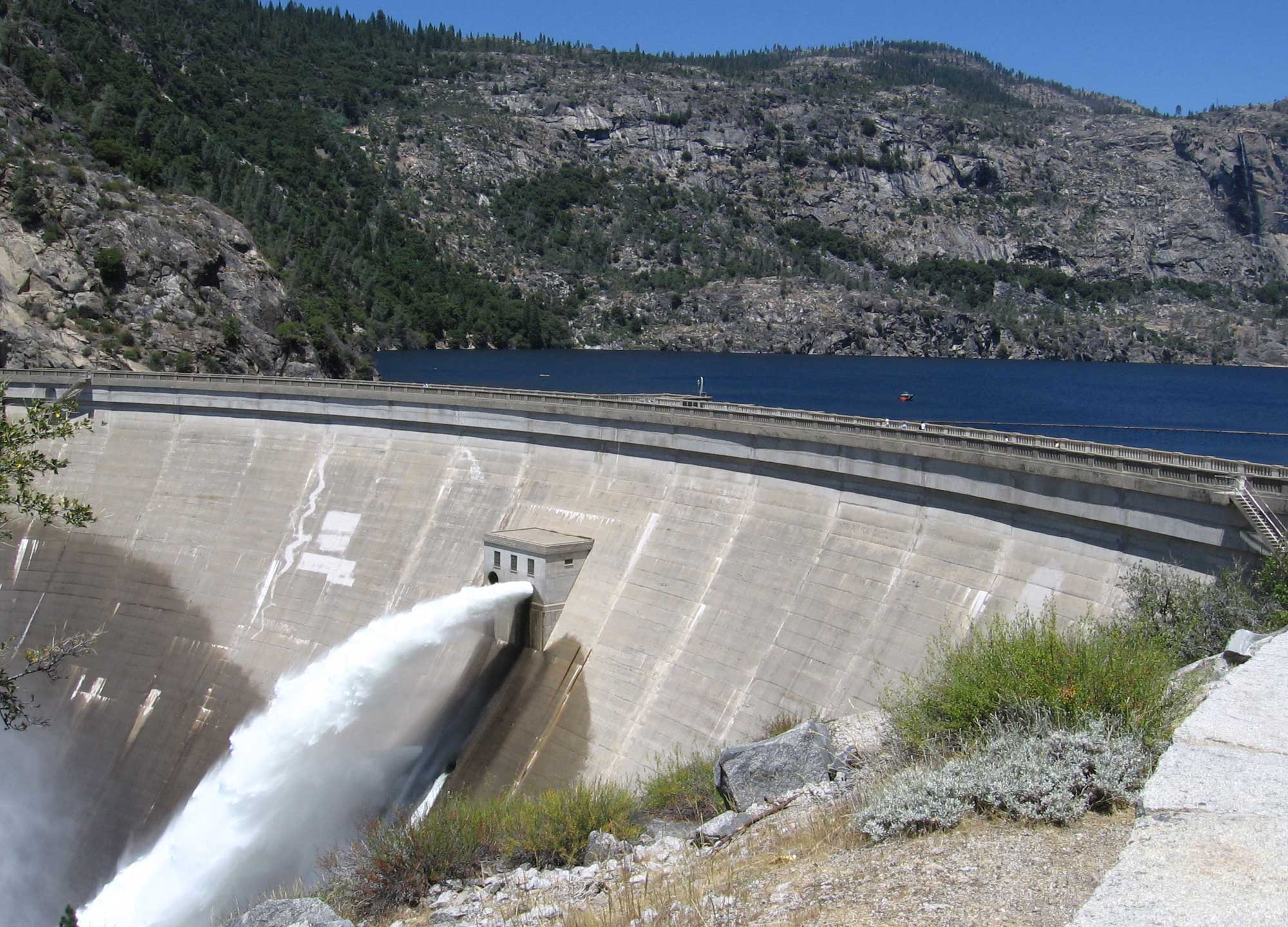 Photograph of O'Shaughnessy Dam in Hetch Hetchy Valley, Yosemite National Park, California. The photo shows a large concrete dam holding back water in a reservoir with rock slopes rising in the background. Water is shooting out of a pipe on the downriver side of the dam.