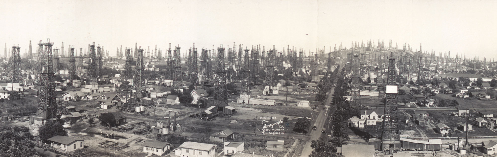 Black and white photograph showing the landscape of Signal Hill, Long Beach, California, ca. 1923. The photo shows numerous oil derricks scattered among buildings which appear to be residential homes.