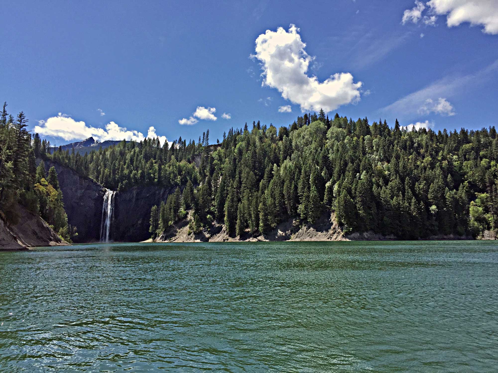 Photograph of PeeWee Falls on Boundary Reservoir, northeastern Washington. The photo shows the surface of a lake in the foreground with conifer-covered hills rising in the background. A waterfall is flowering over a cliff at the far end of the lake.