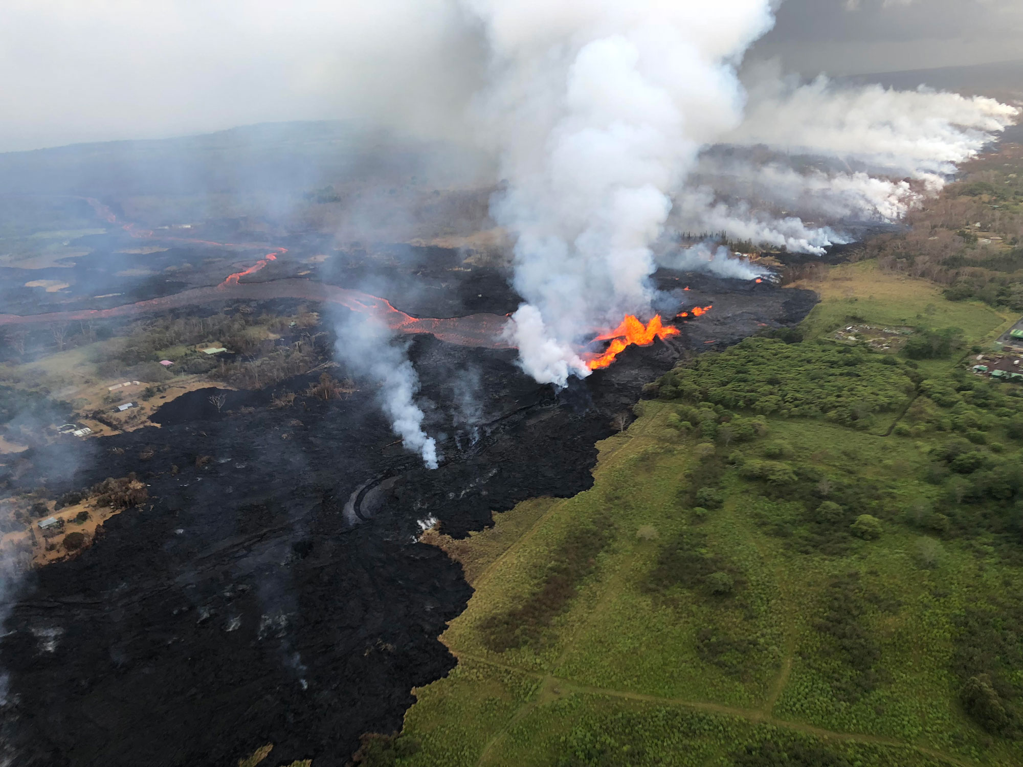 Photograph of a lava flow on Hawai'i in 2018. The lava flow goes from the upper right to lower left of the image. In the background (upper right), smoke rises from the lava flow. In the center of the image, orange lava is visible flowing off to the left center and upper left portions of the image. In the lower left, cooling black lava rock is present. To the right of the lava flow, a green, vegetated landscape is present. At center-right, buildings and infrastructure of the geothermal plant can be seen.