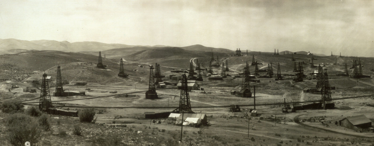 Black and white photograph of the San Francisco oil field ca. 1909. The photo shows a landscape of low-rolling hills. In the foreground is a developed area with multiple oil derricks.
