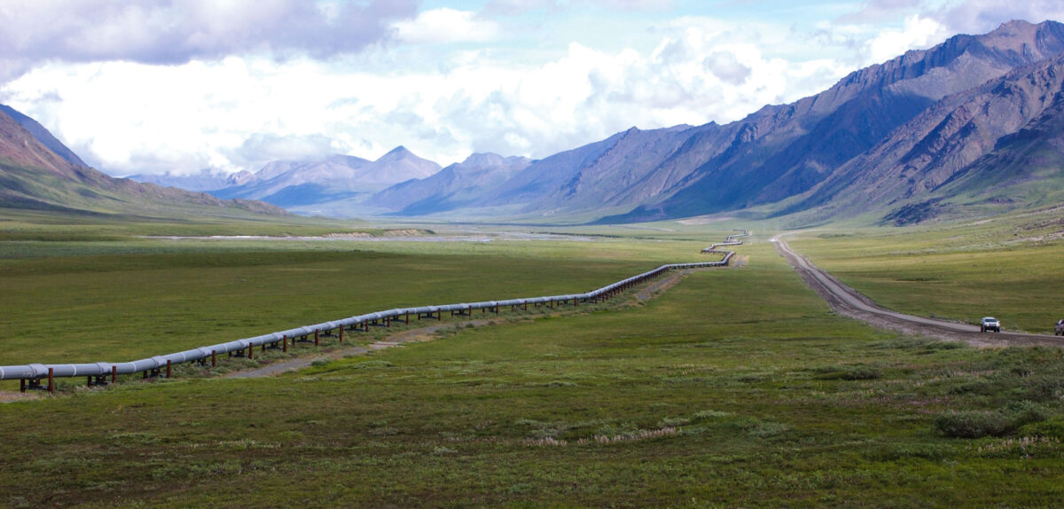 Photograph of the Trans-Alaska pipeline. The photo shows a flat valley with the pipeline running from the right background to the left foreground of the image. A road with two cars on it can be seen to the right of the pipeline. Mountains rise on the right in the background.