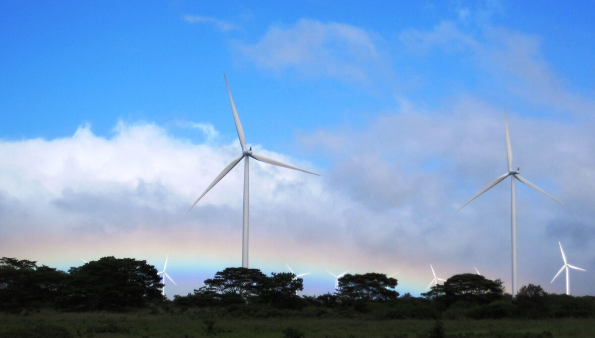 Photograph of large white wind turbines rising high above the treetops in Hawaii. Two turbines are in the foreground behind the trees, more are in the background. A rainbow is also visible just above the treetops. White and gray clouds are scattered across the sky.