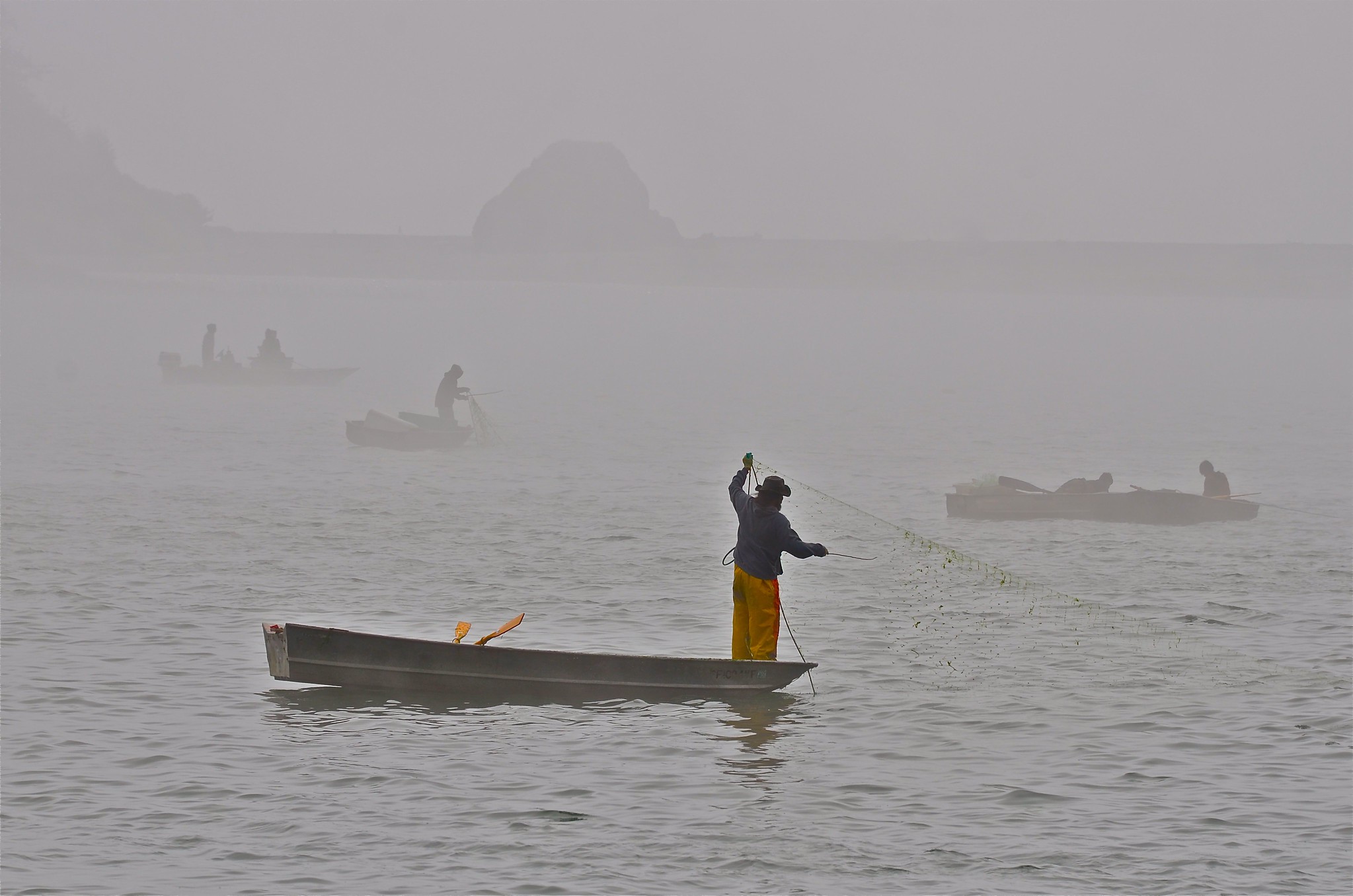 Photograph of Yurok people fishing in the Klamath River. The photos shows small boats (canoes or rowboats) on a river. The nearest one has a man standing in the bow with a large net. In the background, other boats have one or two people sitting or standing.
