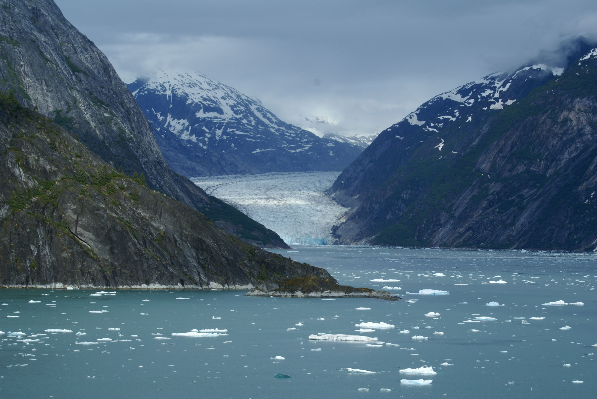 Photograph showing the Dawes Glacier flowing into the water of the Endicott Arm Fjord in Alaska. The photo shows a glacier filling a narrow valley between mountains. The glacier ends at the surface of the water in the fjord, with also has slopes rising to either side.
