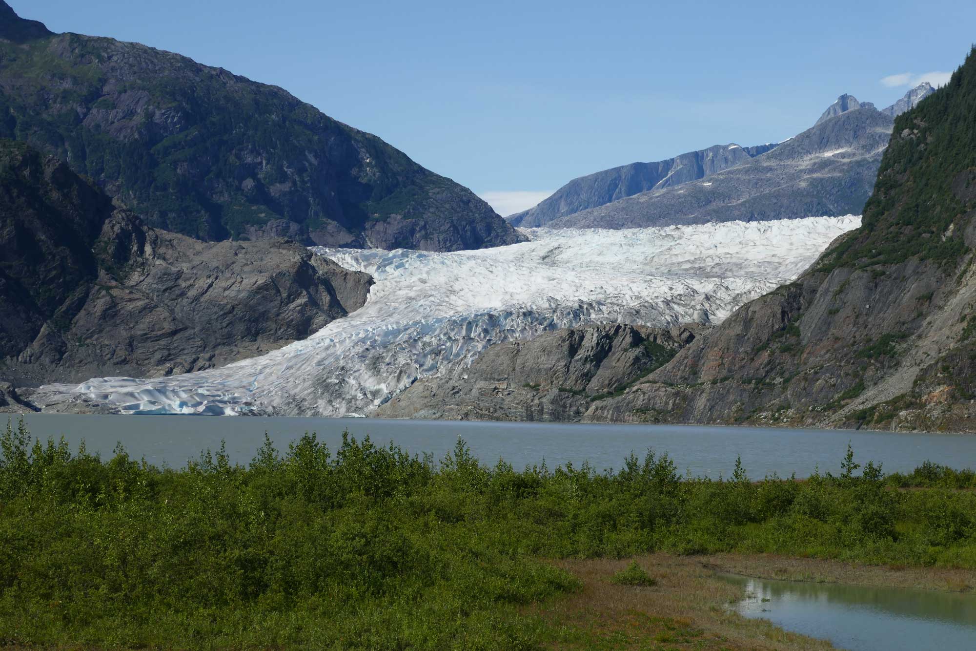Photograph of Mendenhall Glacier, southeastern Alaska. The photo shows a glacier flowing down a rocky slope and into a lake. Vegetation grows on the shore opposite the glacier.