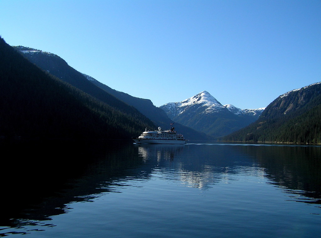 Photograph of Misty Fjords National Monument in Alaska. The photo shows a large ship in an arm of the sea at the base of sloping hillsides covered with conifers. A snow-capped mountain peak rises in the background.