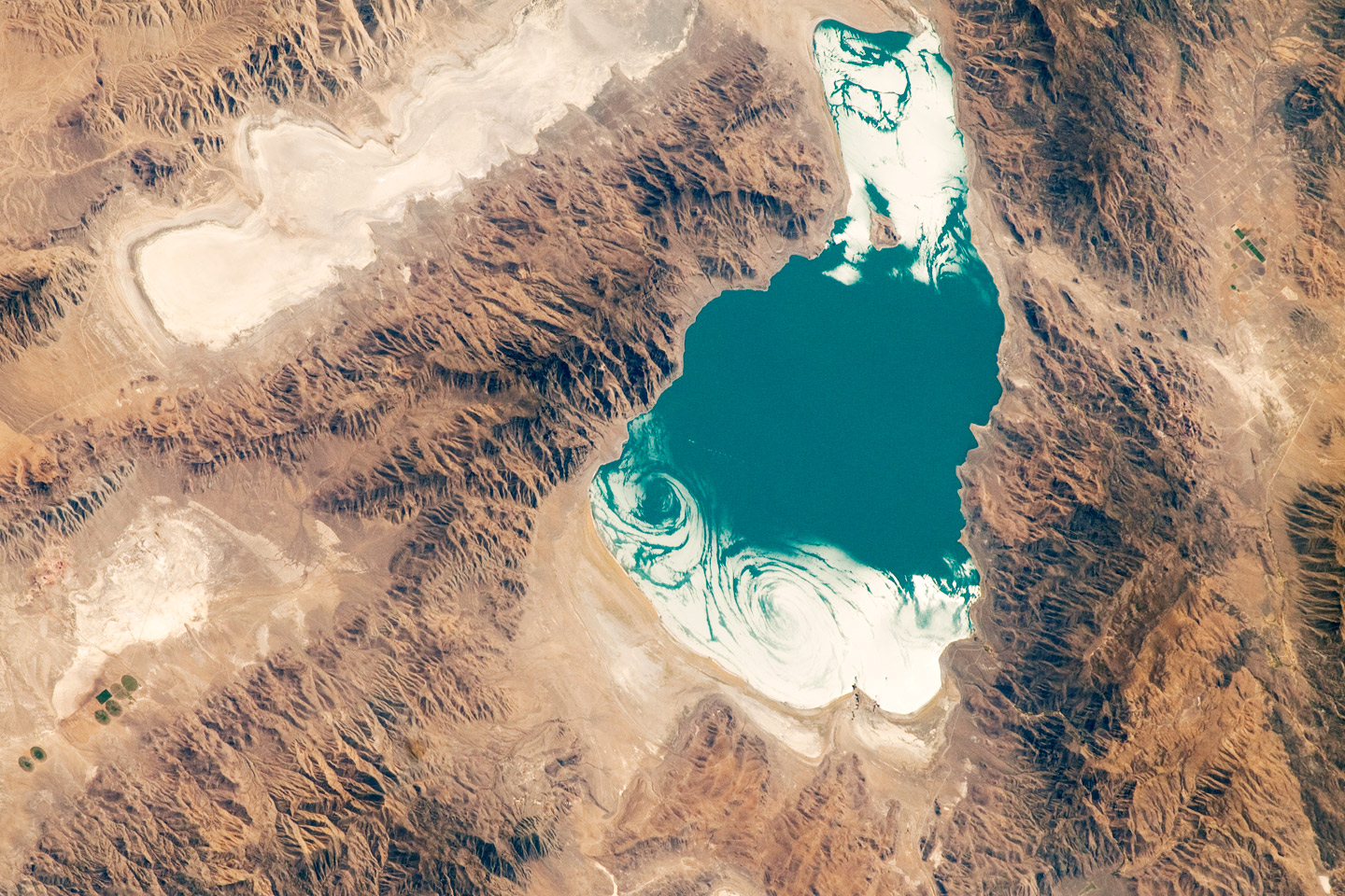 Satellite photo of Pyramid Lake, Nevada. The photo shows an irregularly-shaped lake at center right; part of the lake surface is blue, but but ends have swirls of white. Surrounding the lake, the landscape has a series of mountains ranges separated by dry basins. A few small, circular patches of green irrigated land can be see at the lower left.