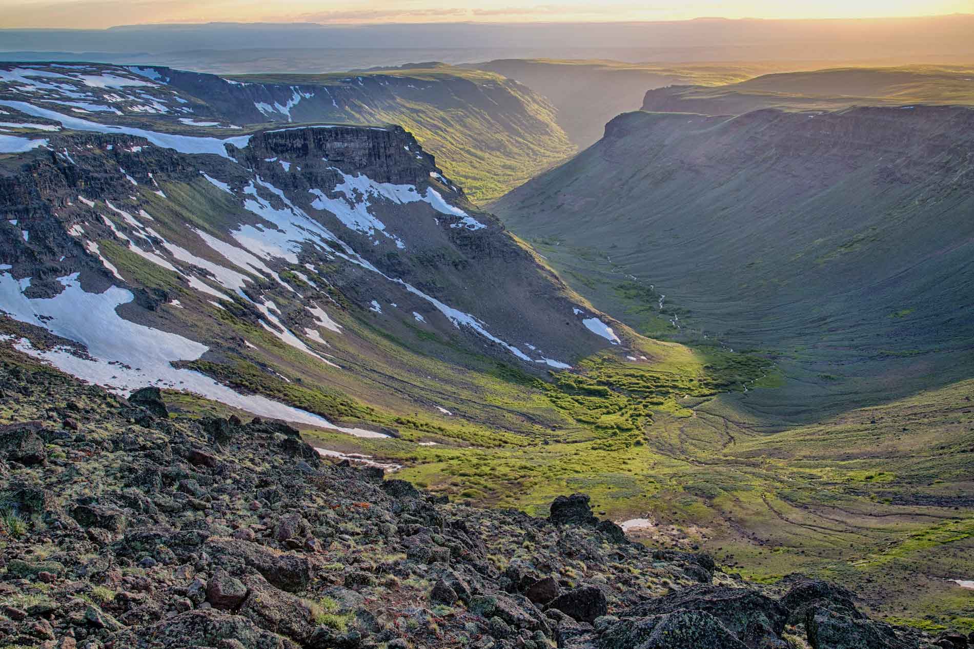 Photograph of a U-shaped glacial valley on Steen Mountain, Oregon. The photo shows a valley with a broad, U-shaped floor snaking between flat-topped hills. The valley floor has green vegetation and the hillsides are covered in patches of white snow.