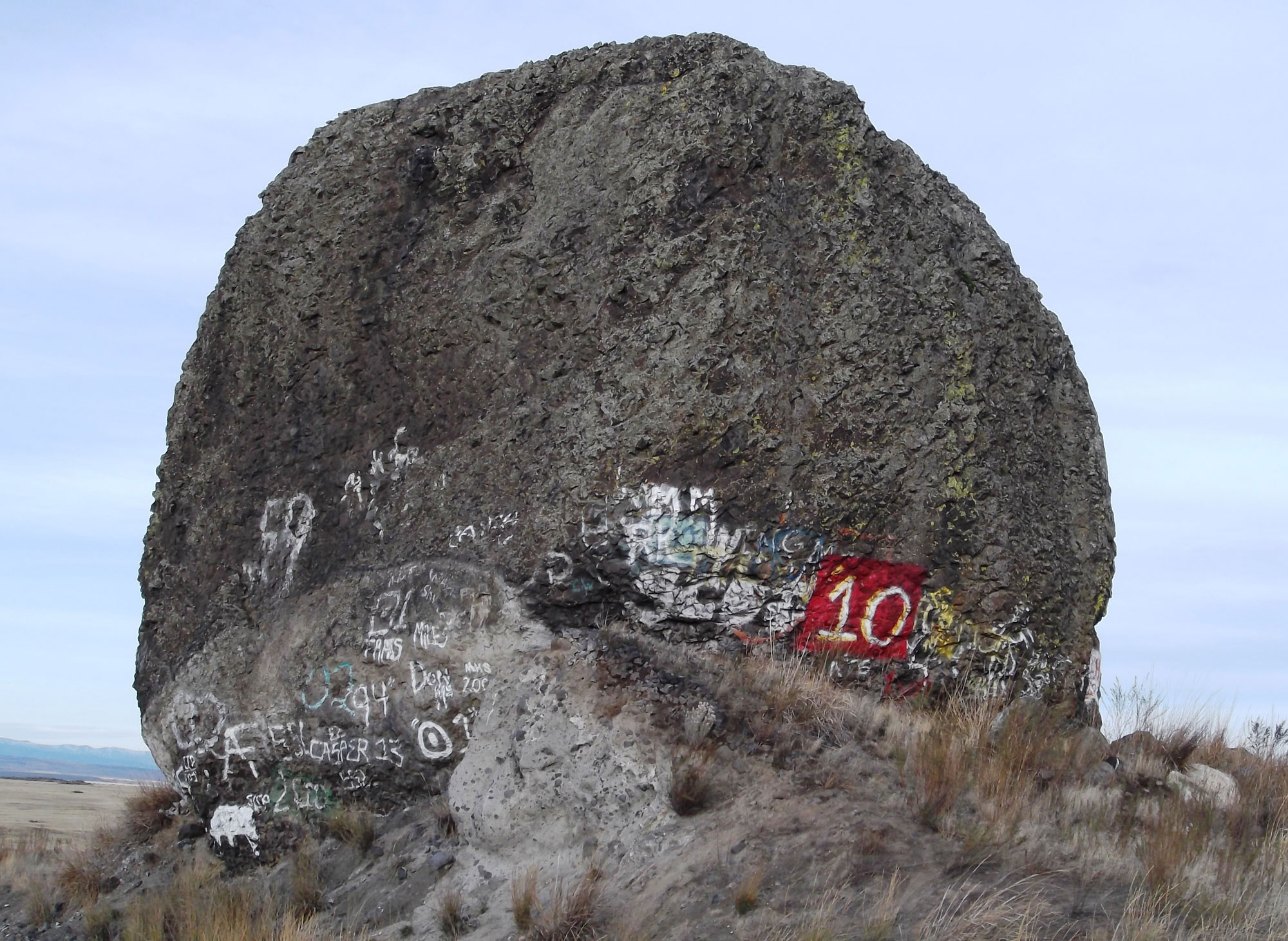 Photo of Yeager rock, a large glacial erratic in Washington. The photo shows a large, round, gray boulder sitting on a small hill covered with dry grass. The lower part of the boulder is covered with graffiti.