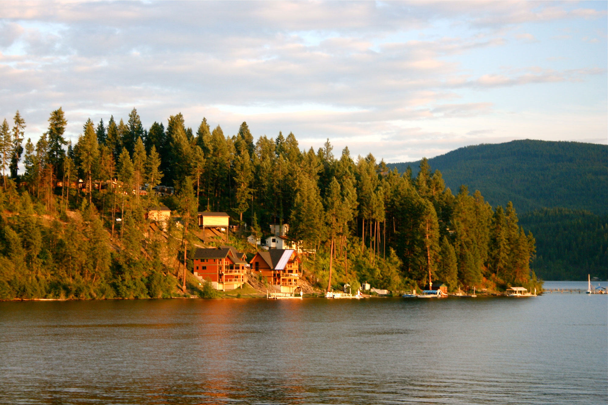 Photograph of Coeur d'Alene, Idaho. The photo shows a lake in the foreground with conifer-covered hills rising at its edges. Houses with docks and boats can be seen near the lakeshore in the middle ground of the image.