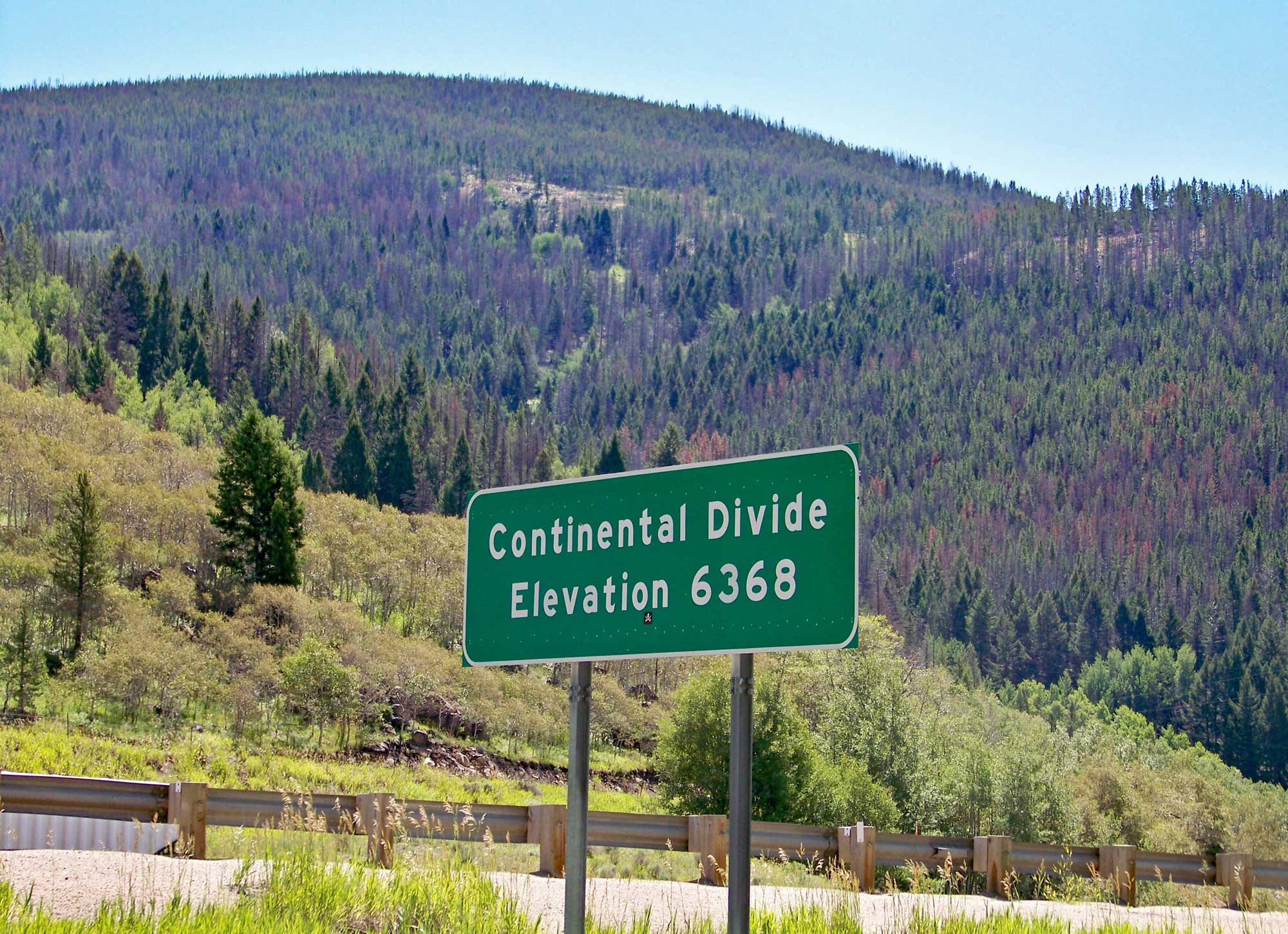 Photograph of the continental divide at Elk Park Pass, Montana, in 2010. The photo shows a green and white road sign that says "Continental Divide, Elevation 6368" in the foreground in front of a highway guardrail. Hills covered with conifers and broadleaved trees rise in the background.