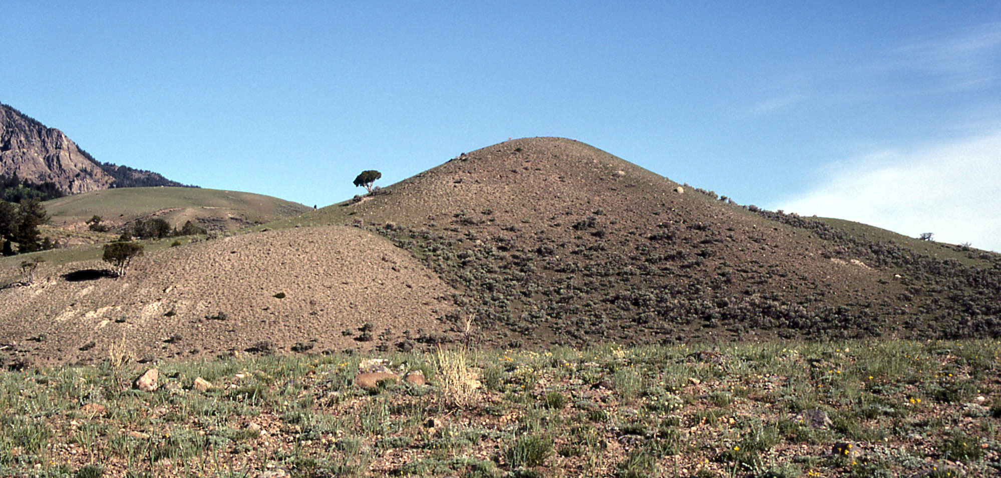 Photograph of Dude Hill, Yellowstone National Park. The photo shows a low, rounded hill that is sparsely vegetated. A flat landscape with tufts of grass occurs in the foreground. In the left background, another hill and a rocky cliff rise.