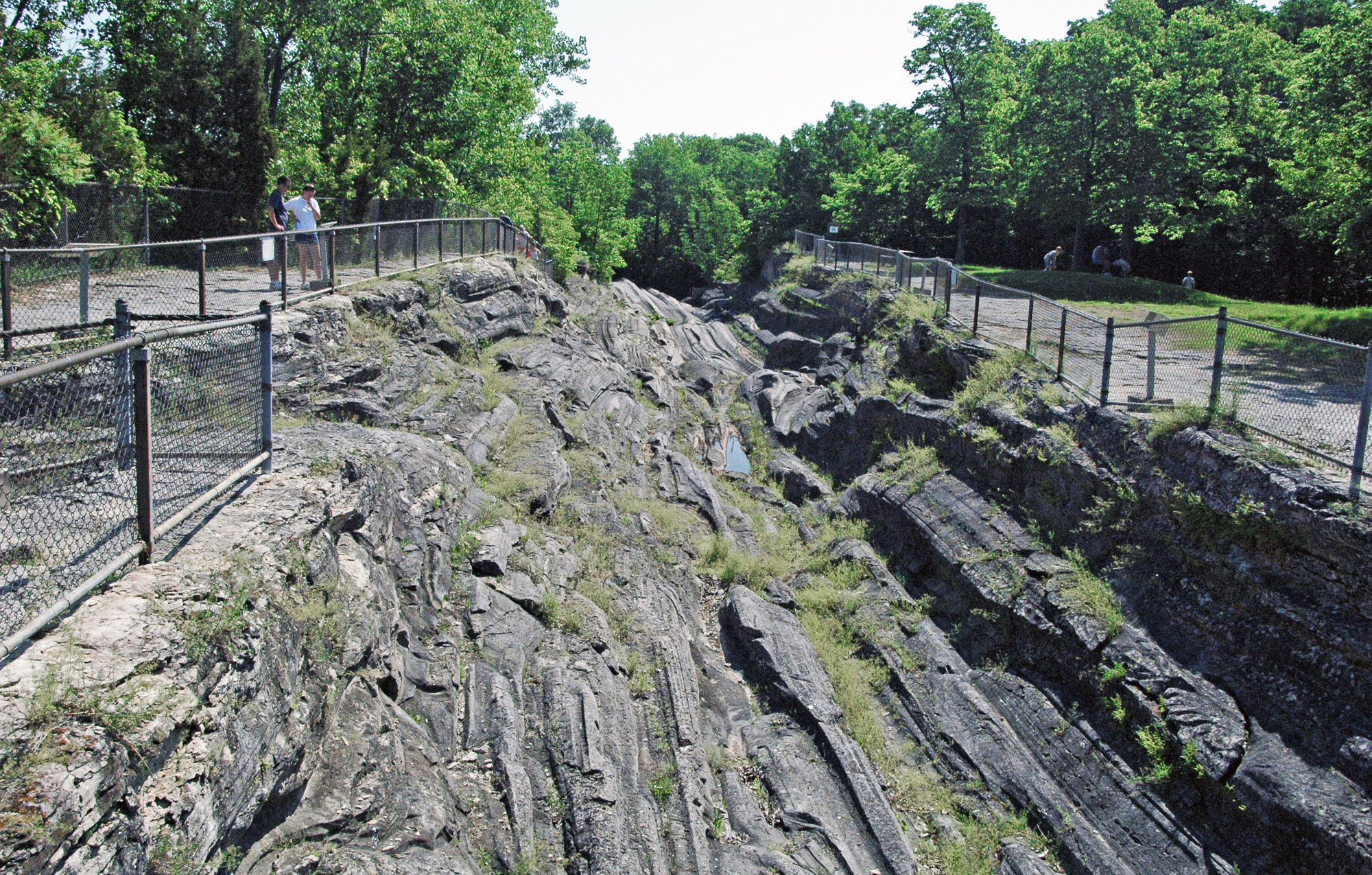 Photograph showing glacial grooves on Kelleys Island in Lake Erie, Ohio. The photo shows a linear channel carved through gray rock. The channel is lined with long, roughly parallel grooves. A chainlink fence borders either side of the channel, and two people can be seen behind the fence on the left of the image looking into the channel. Around the channel, the landscape is forested.